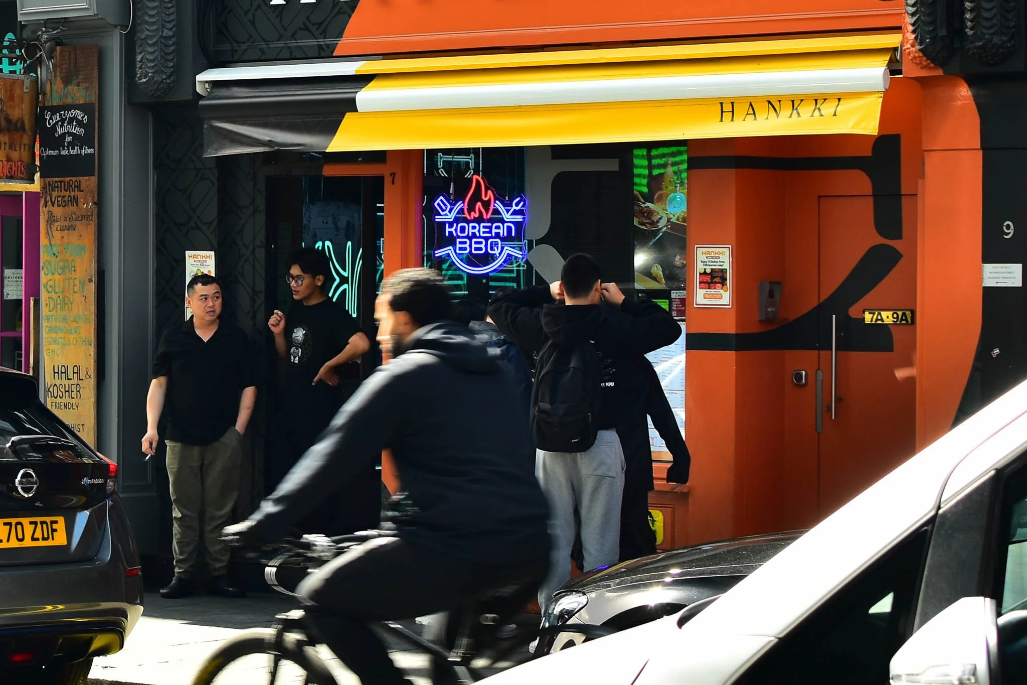 Street scene outside a Korean barbecue restaurant with people standing and talking, a cyclist passing by, and cars parked along the street.