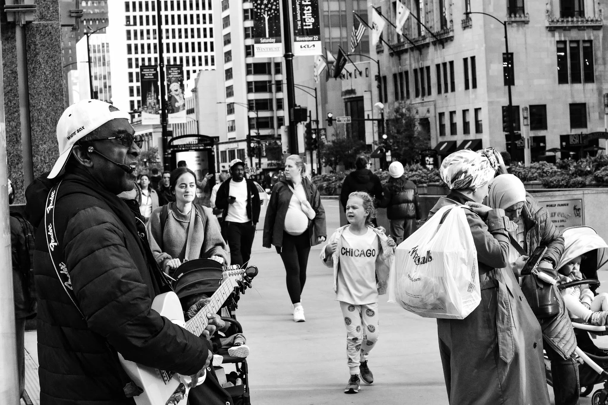 Street scene in Chicago with diverse pedestrians including a man playing guitar, children, and adults, with tall buildings and city signage in the background.