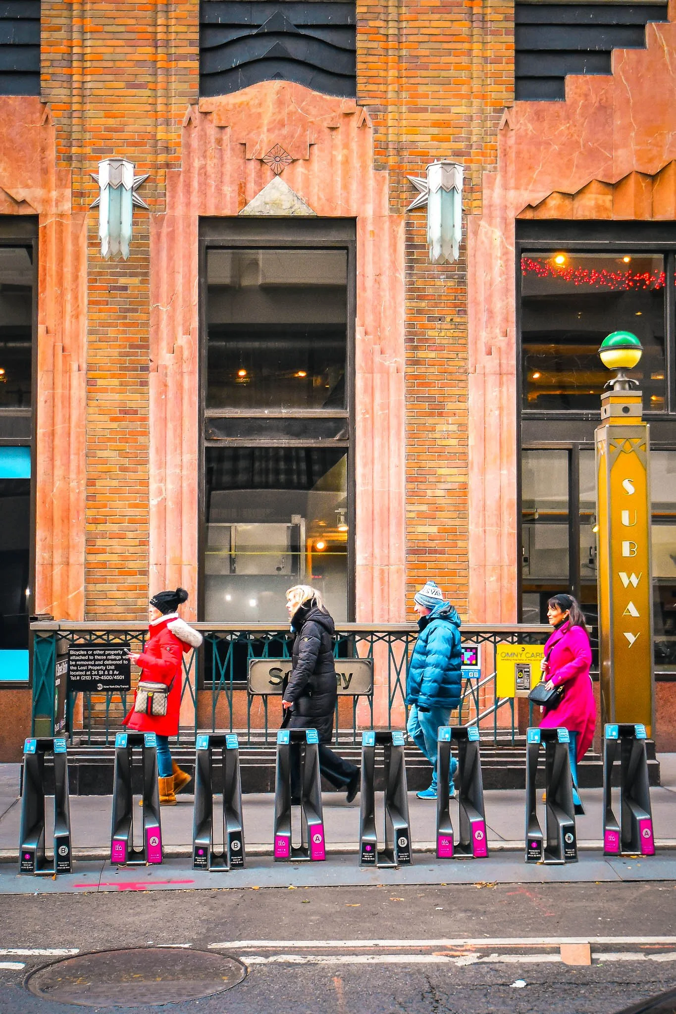 People standing in line outside a subway station in an urban setting, with a brick building in the background.
