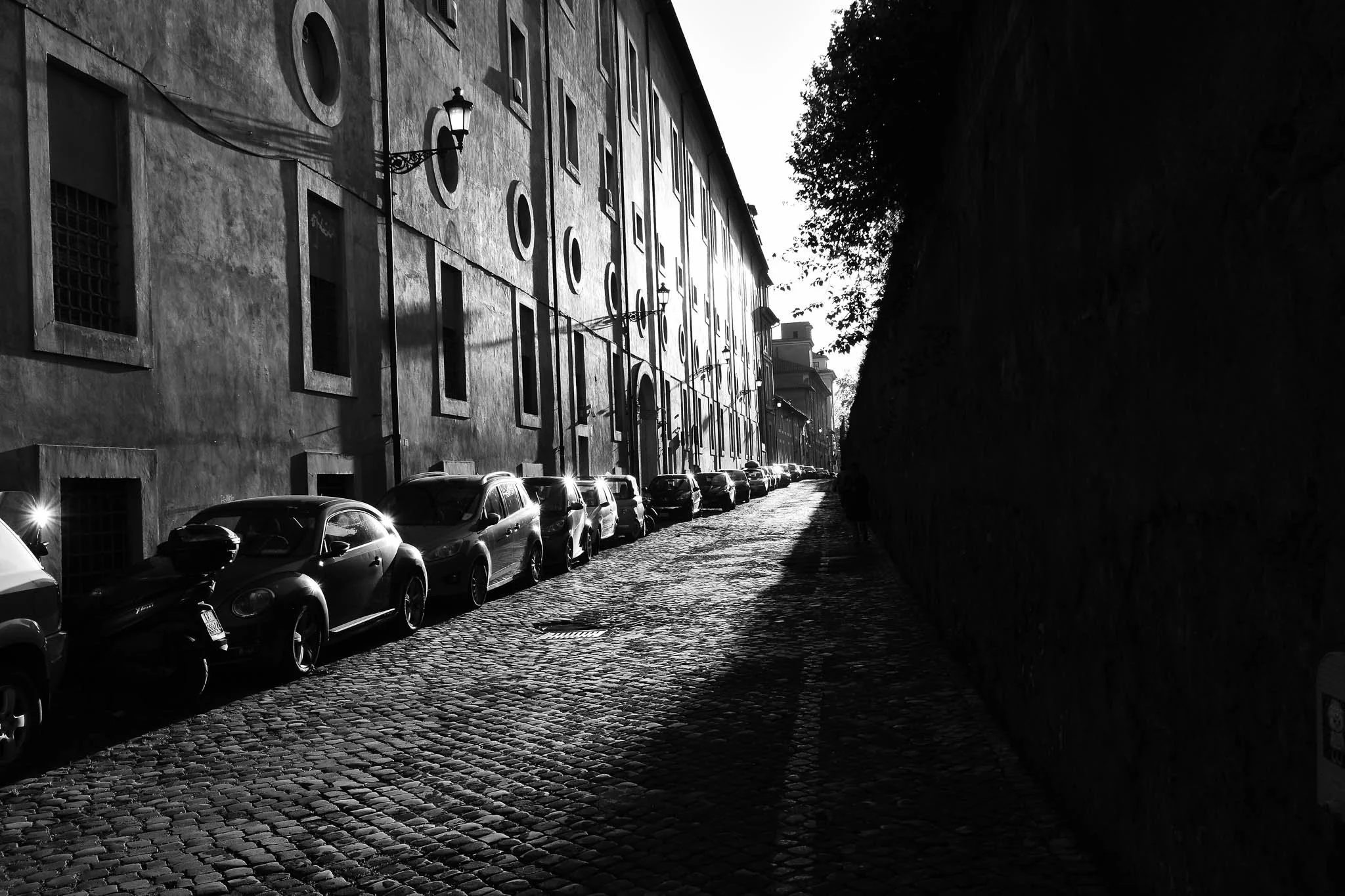 Black and white photo of a cobblestone street with parked cars on the left side and a tall building with circular and rectangular windows on the left. A street lamp is mounted on the building, and a large dark wall runs along the right side of the st