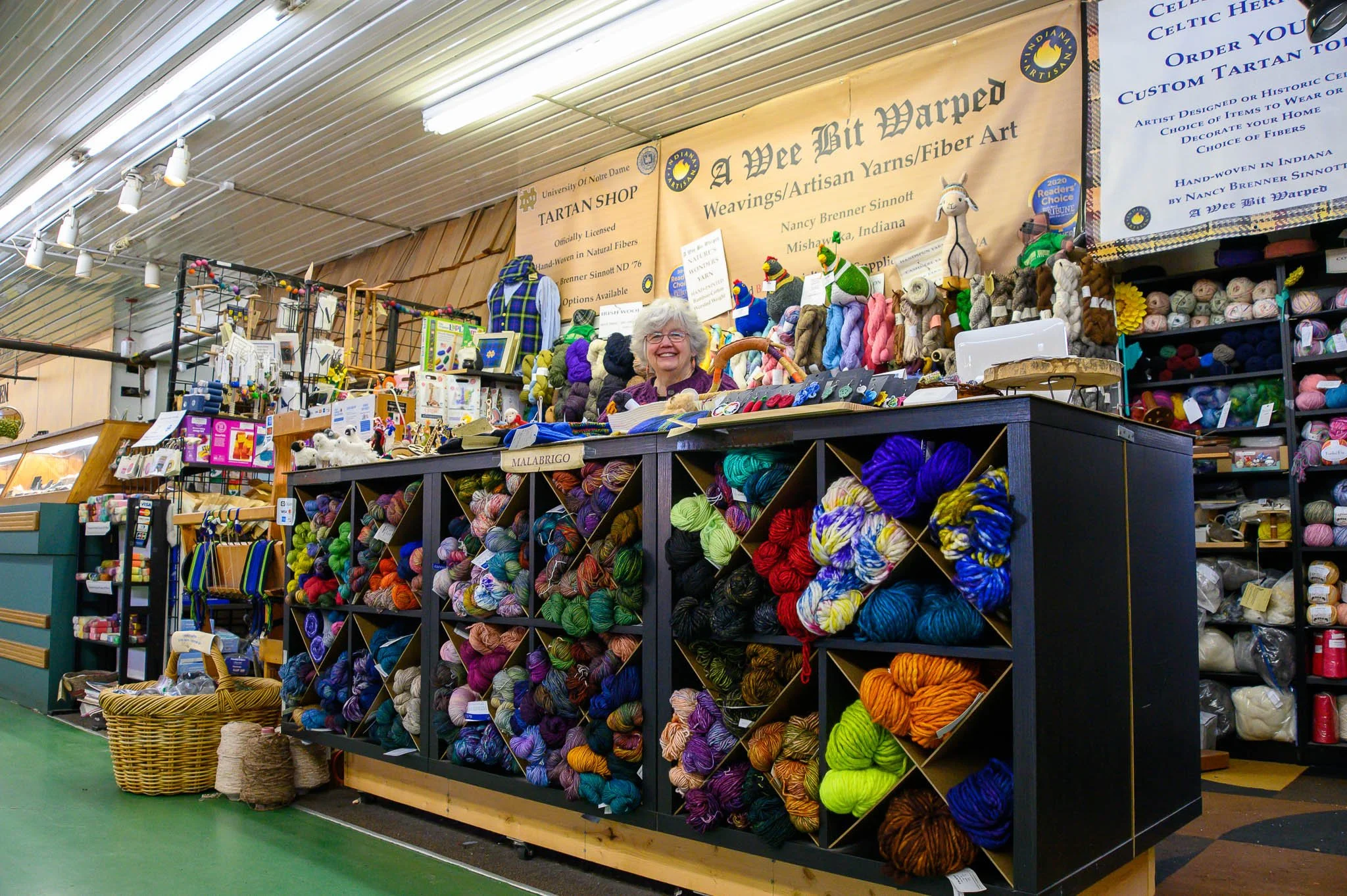 A woman standing behind a display of colorful yarn at a craft fair. She is smiling and surrounded by handmade yarn crafts and accessories.