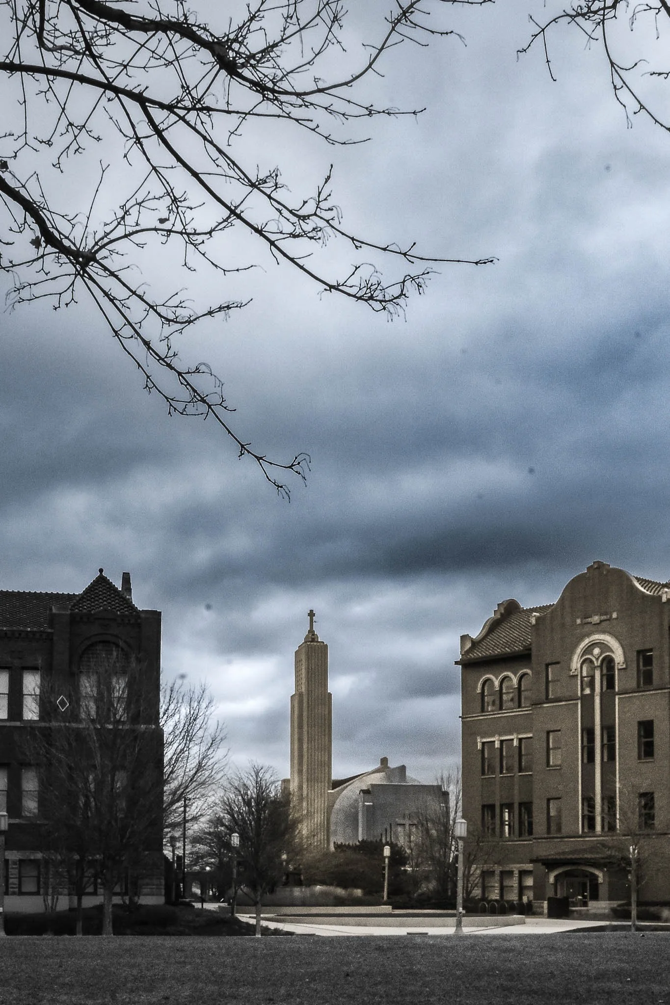 Cityscape with historic buildings, a tall church steeple, trees, and a cloudy sky.