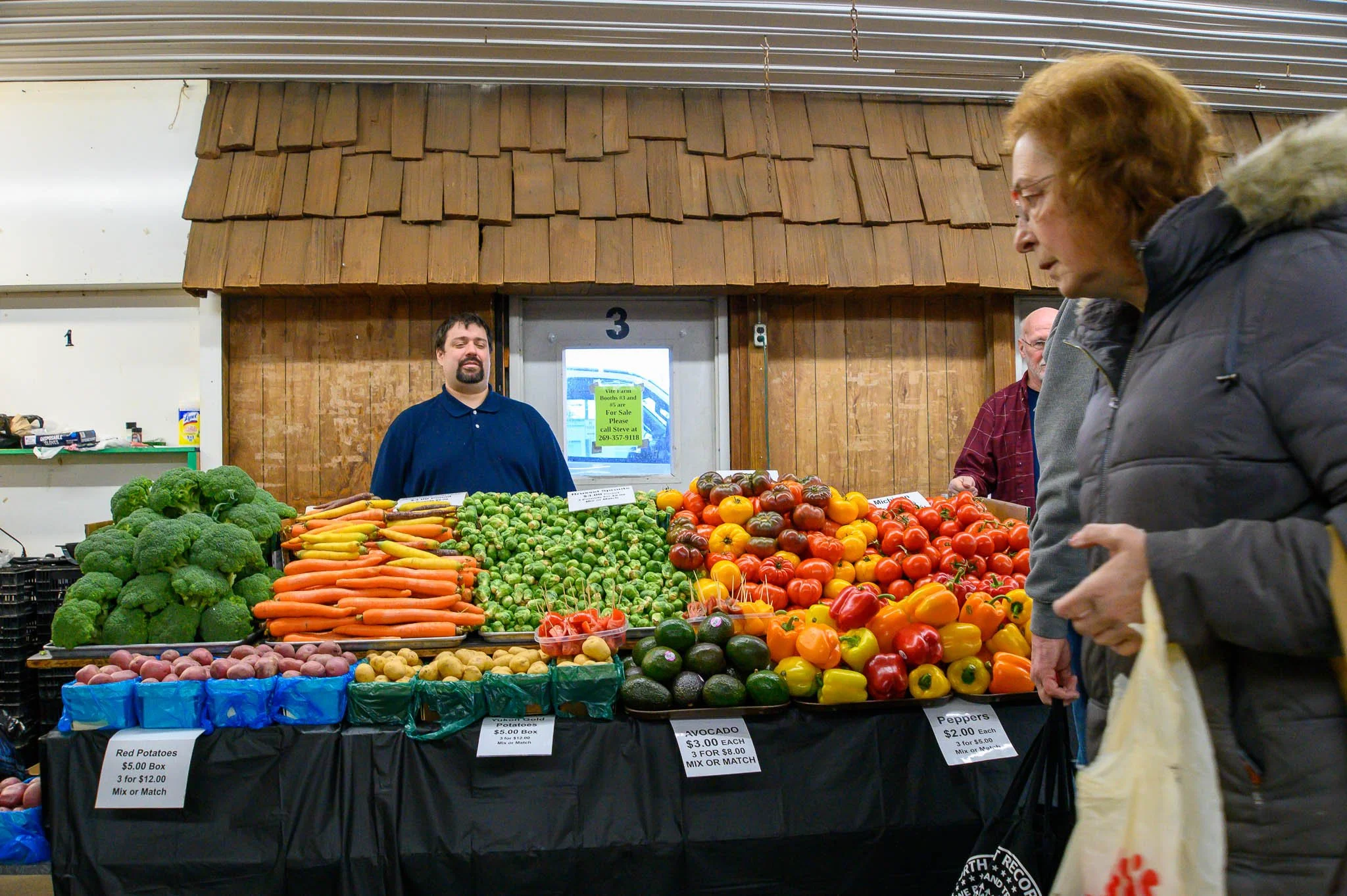 Farmer's market stand with various fresh vegetables including broccoli, carrots, Brussels sprouts, potatoes, avocados, bell peppers, and tomatoes, with customers shopping and sellers attending.