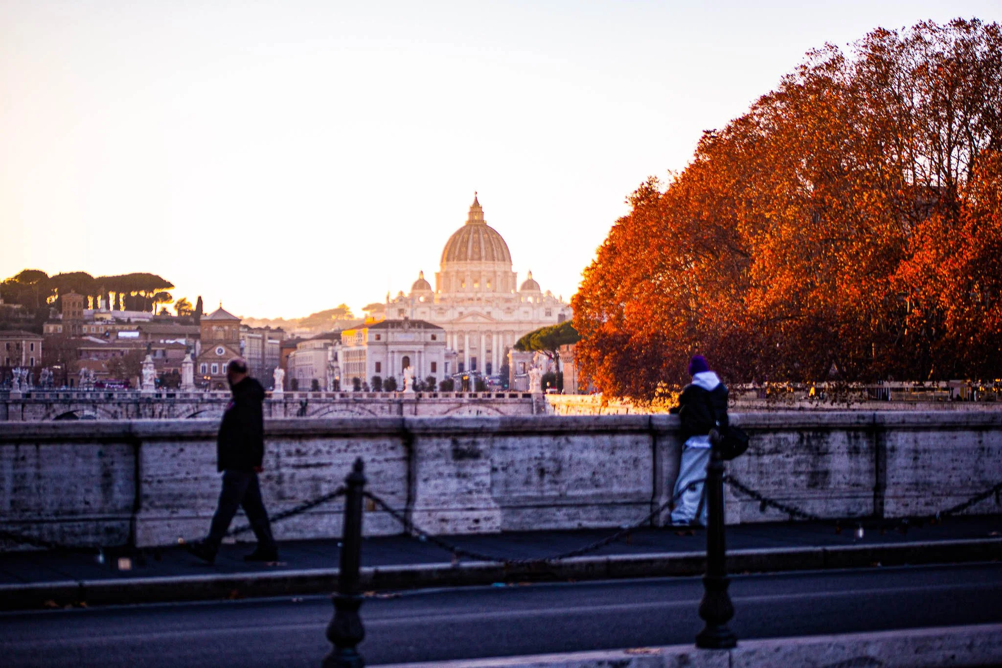 A view of a cityscape featuring a large domed building in the background, with trees and buildings surrounding it. Two people are walking on the sidewalk in the foreground, one holding a bag and the other standing near a tree with orange leaves. The 