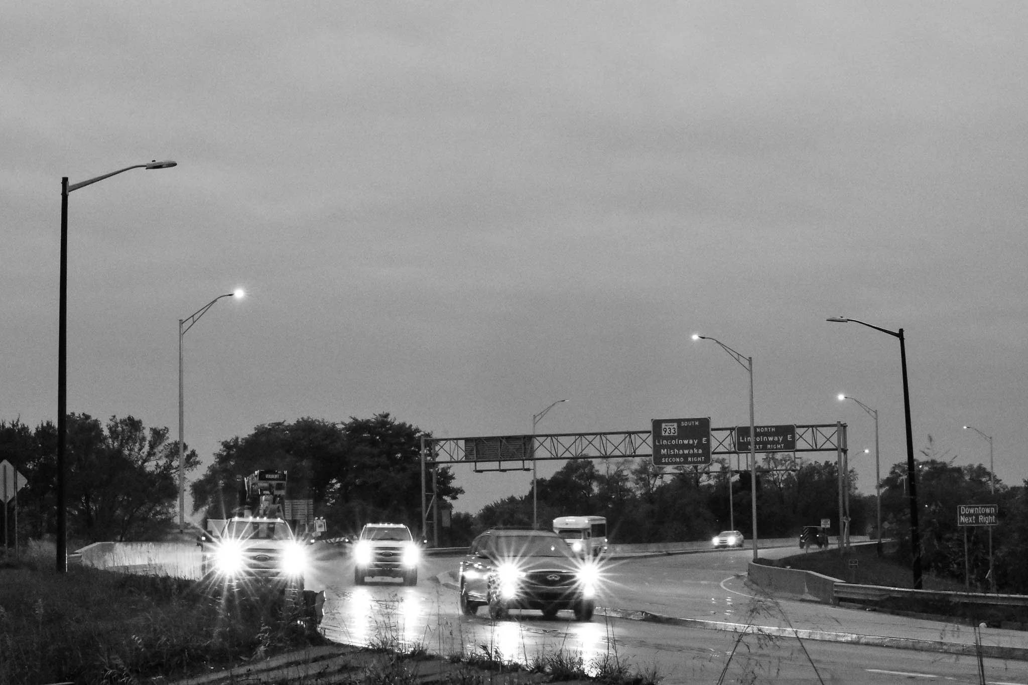 Black and white photo of a highway with cars driving in both directions, streetlights lining the road, and directional signs overhead indicating routes to Lincolnway E, Mishawaka, and Downtown.