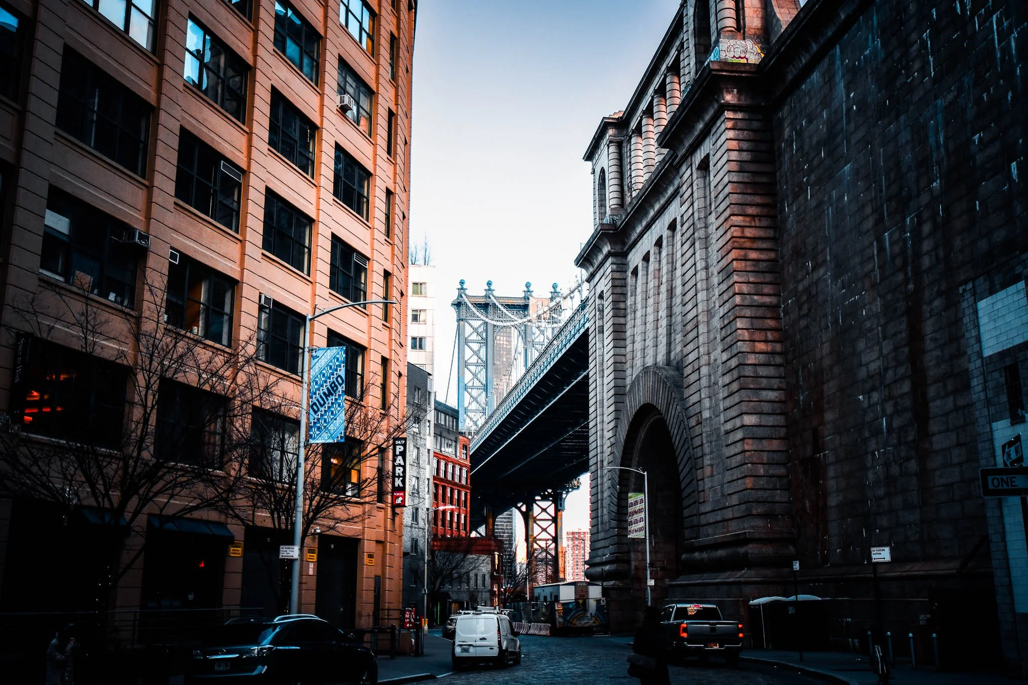 View of a city street with tall buildings, an archway under the Manhattan Bridge, and vehicles parked along the street.