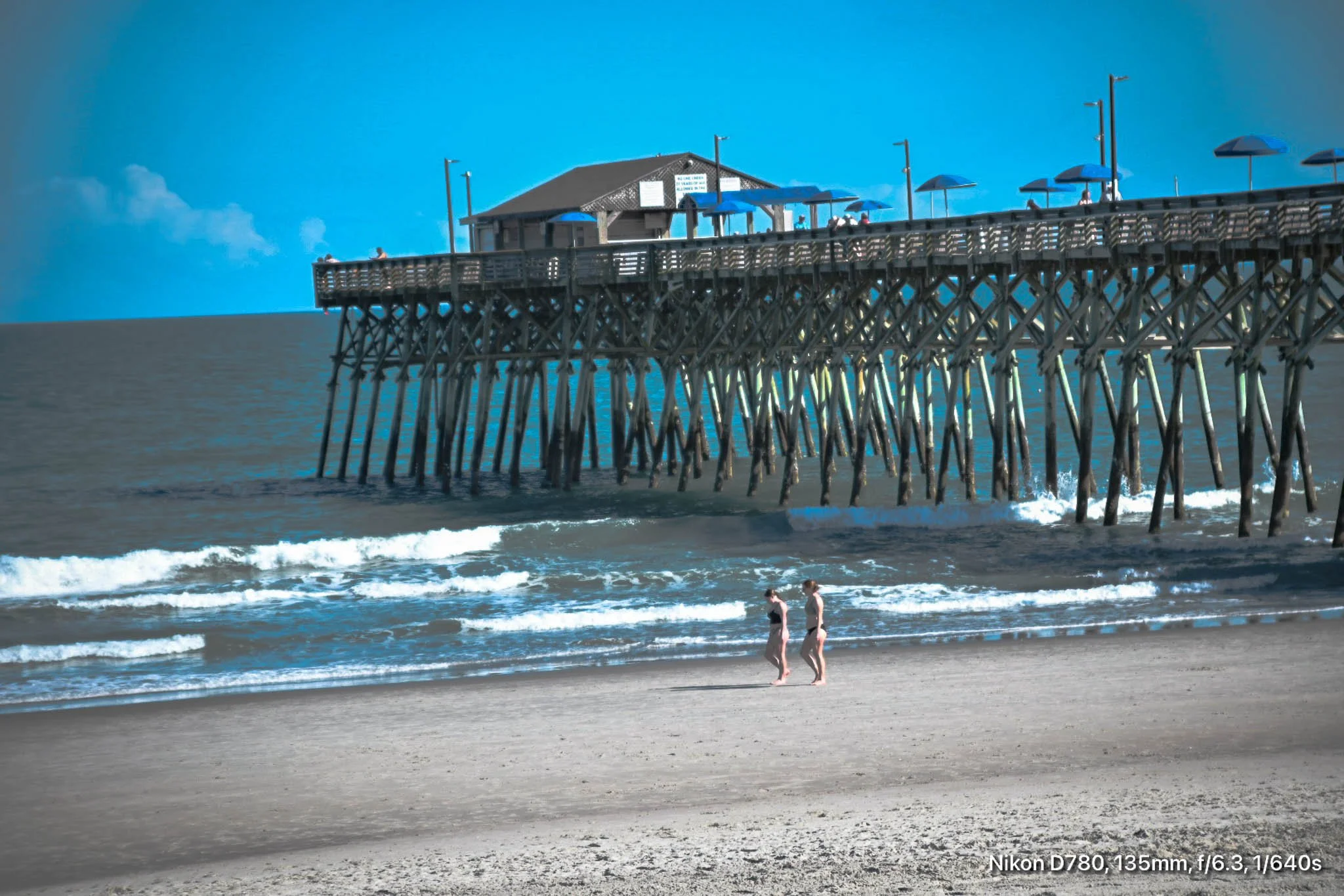 A pier at Garden City, SC extending over the ocean with blue umbrellas on top. Two people walk along the sandy beach below the pier.