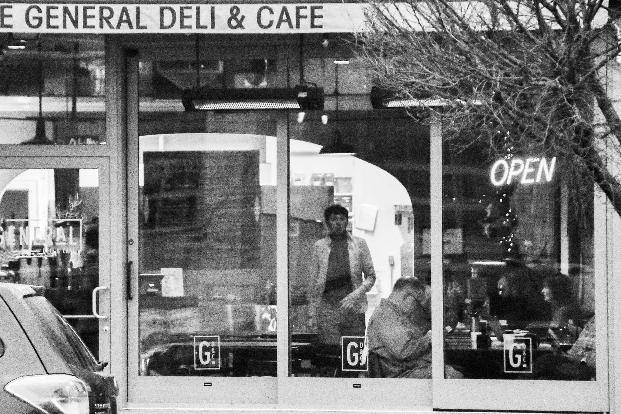 Front view of a deli and cafe with glass windows, showing two people inside, one standing and one sitting. The cafe sign reads 'GENERAL DELI & CAFE', with an illuminated 'OPEN' sign and a tree with bare branches outside.