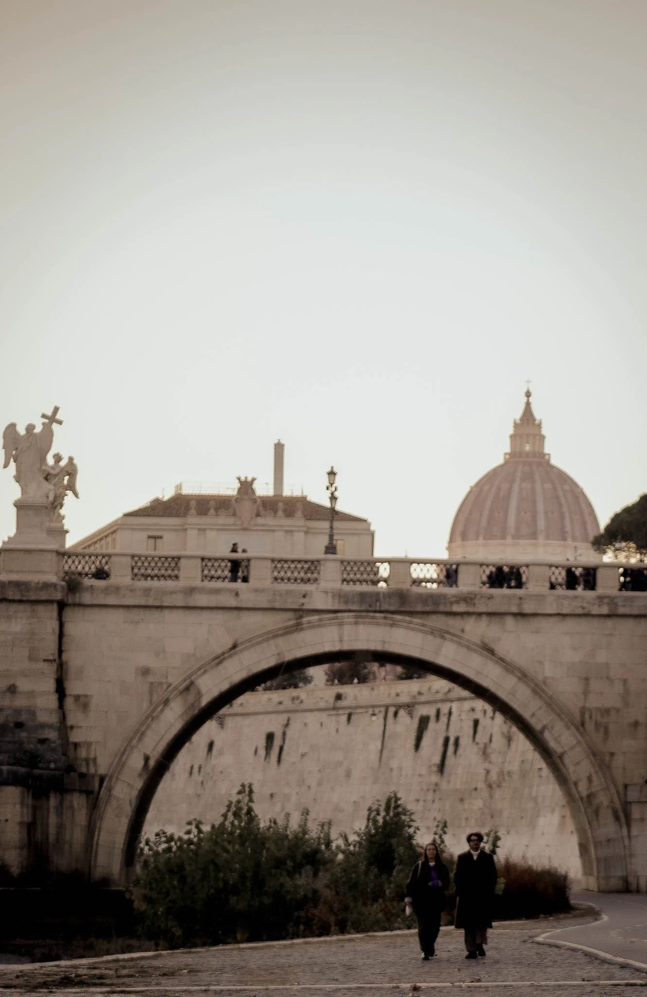 Two people walking under a stone bridge with a large dome in the background, possibly in Rome, Italy. The scene is lit with soft, natural light.
