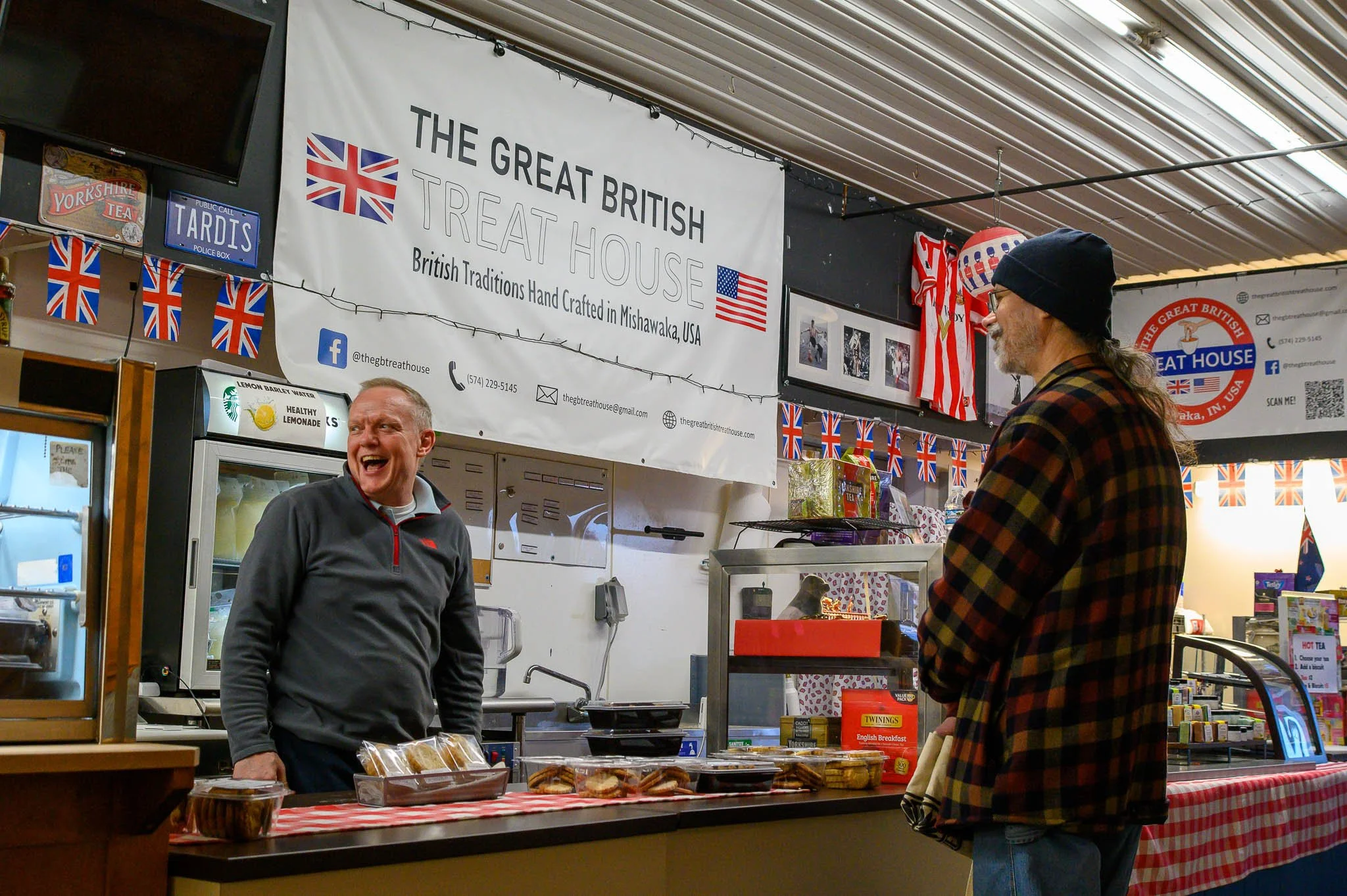 A man with a big smile wearing a gray jacket stands behind a counter at The Great British Treat House, talking to a customer in a checkered jacket and beanie inside a shop decorated with British flags and signs.