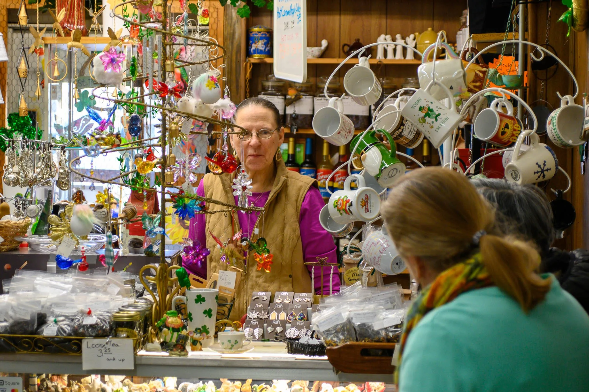 A woman behind a store counter decorated with Christmas ornaments, mugs, and jewelry, with two customers looking at items. The store has wooden shelves and a whiteboard in the background.