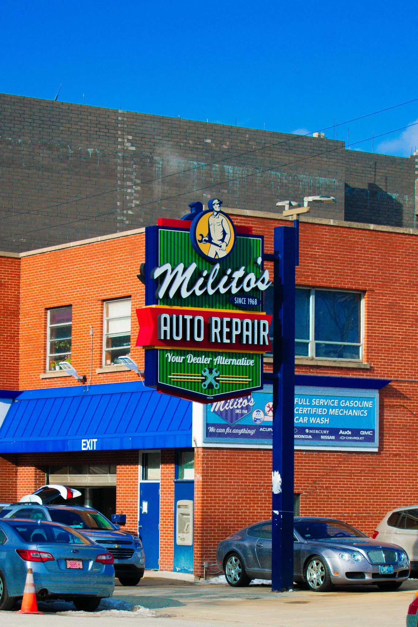 Sign for Militos Auto Repair in front of a red-brick building with cars parked outside.