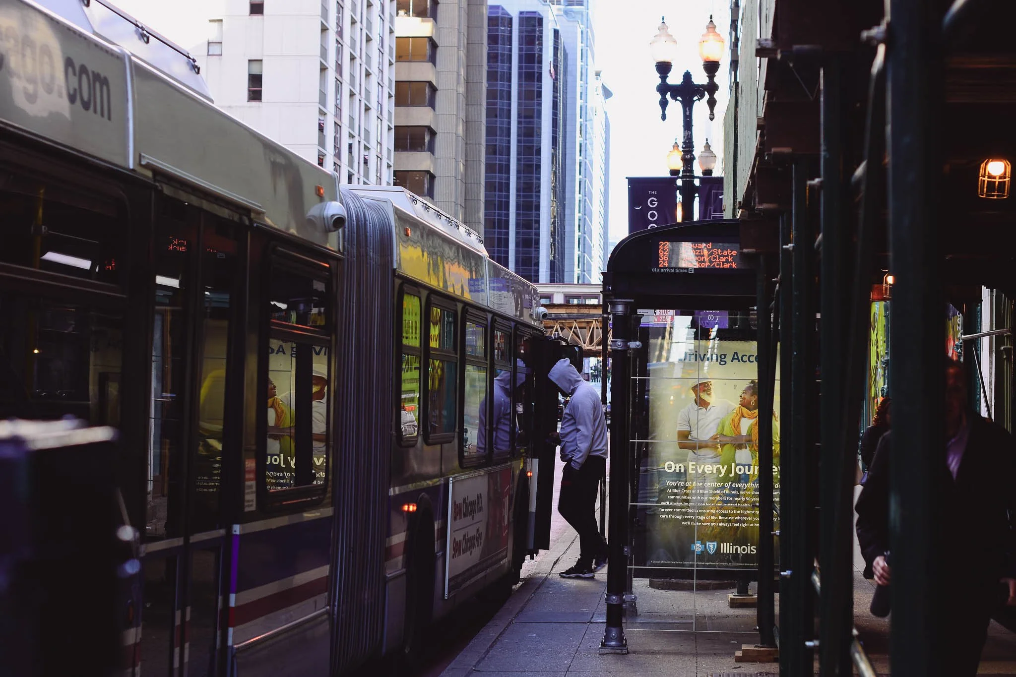 People waiting at a bus stop in an urban cityscape, with tall buildings, street lamps, and bus signage.