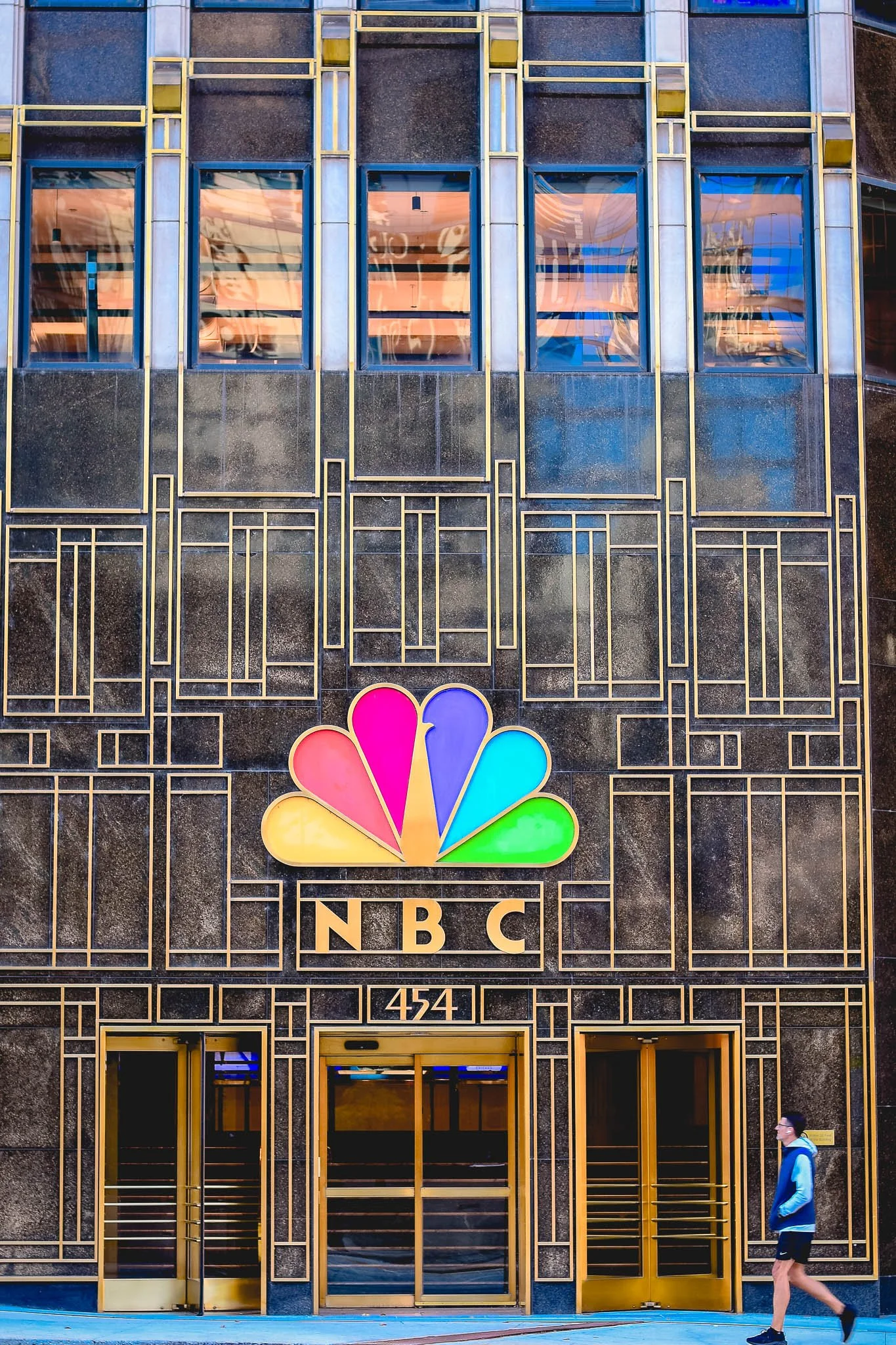 Front of NBC building with large NBC logo and gold accents, a person walking by in casual clothing.