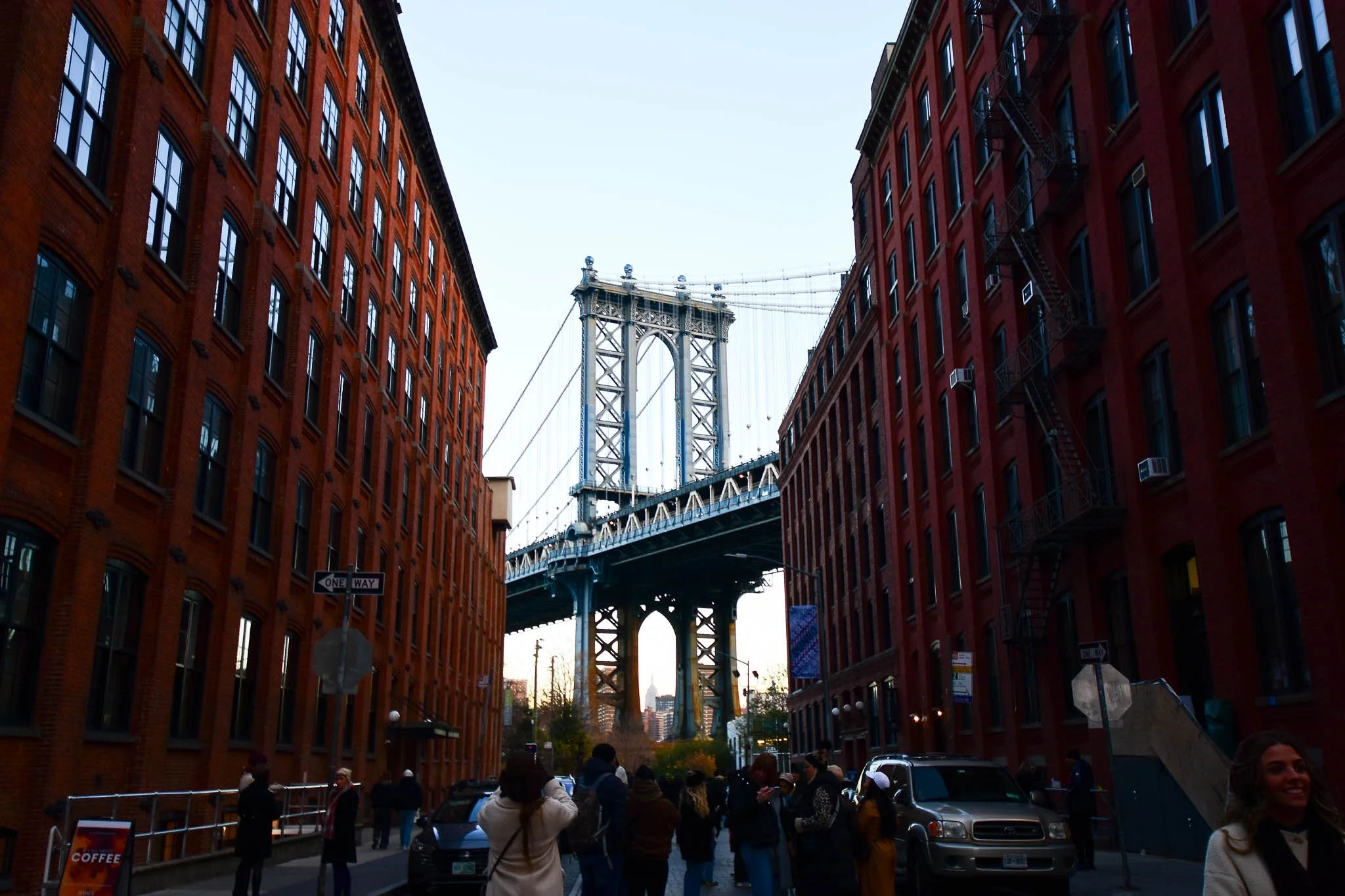 View of the Manhattan Bridge through a city street with people walking and parked cars, buildings on both sides, and a clear sky.