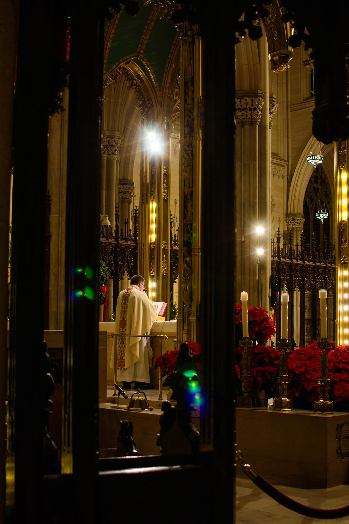A priest standing at an altar inside a church, seen through a decorative barrier with Christmas poinsettias and lit candles, illuminated by warm lighting and behind Gothic-style architecture.