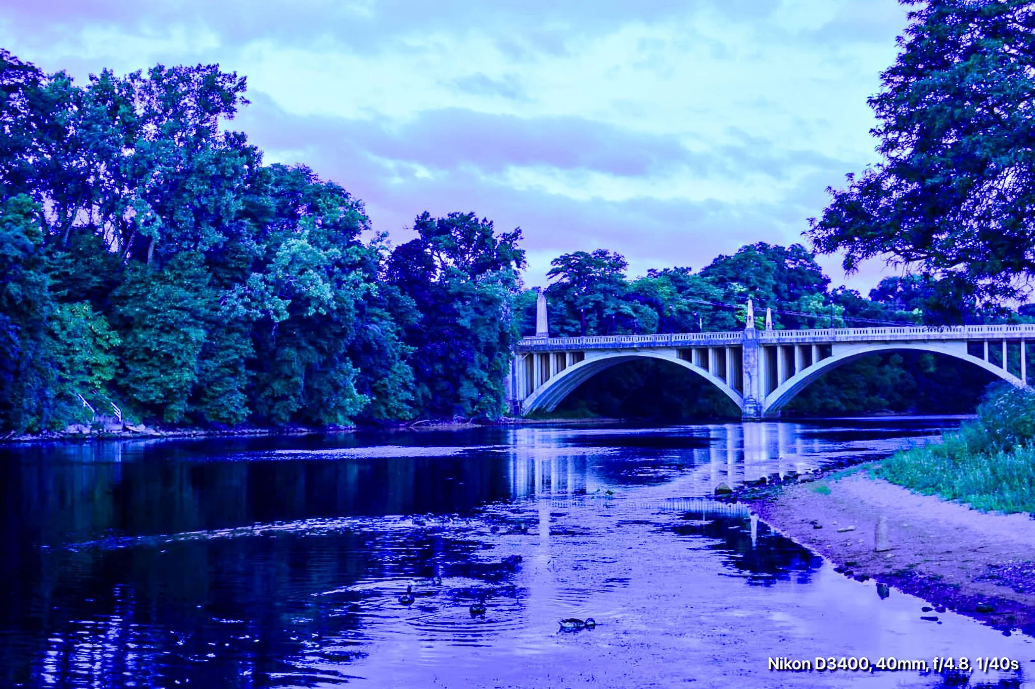 A river with a bridge and lush green trees on both sides, under a cloudy sky.