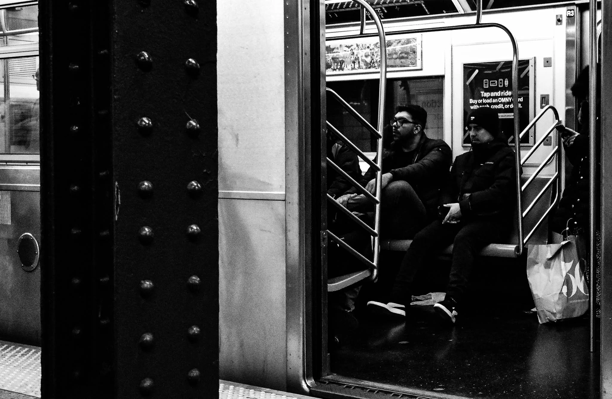 Black and white photo of three people sitting inside a subway train, looking at their phones. The train has metallic seats and railings, with an information display in the background. The photo captures a side view of the open door of the subway car,