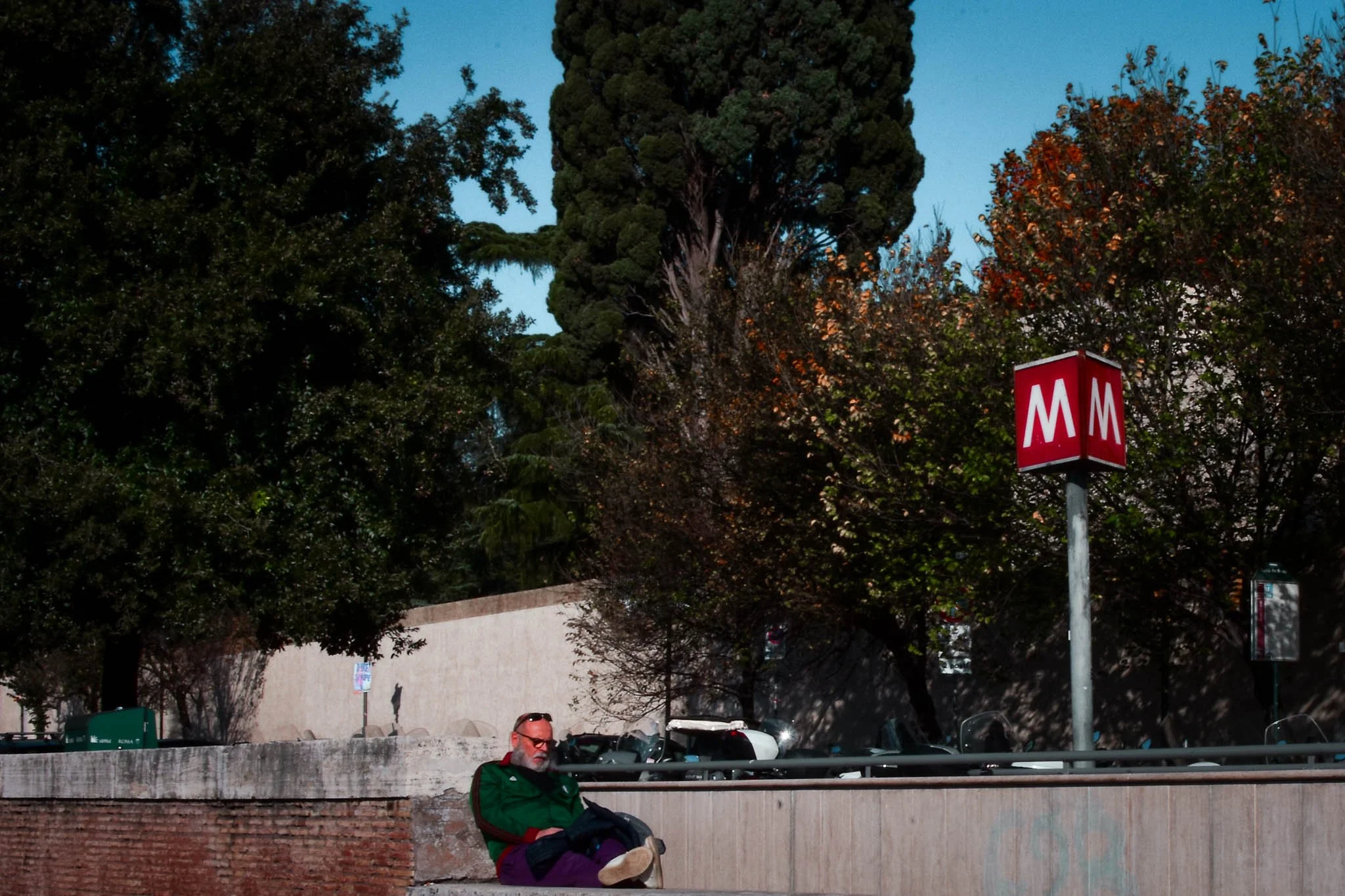 A man with sunglasses and a beard, wearing a green jacket, sits on the sidewalk with his feet up, next to a brick wall and a metal railing. There are trees and cars in the background, and a red sign with white 'M's indicating a metro station.