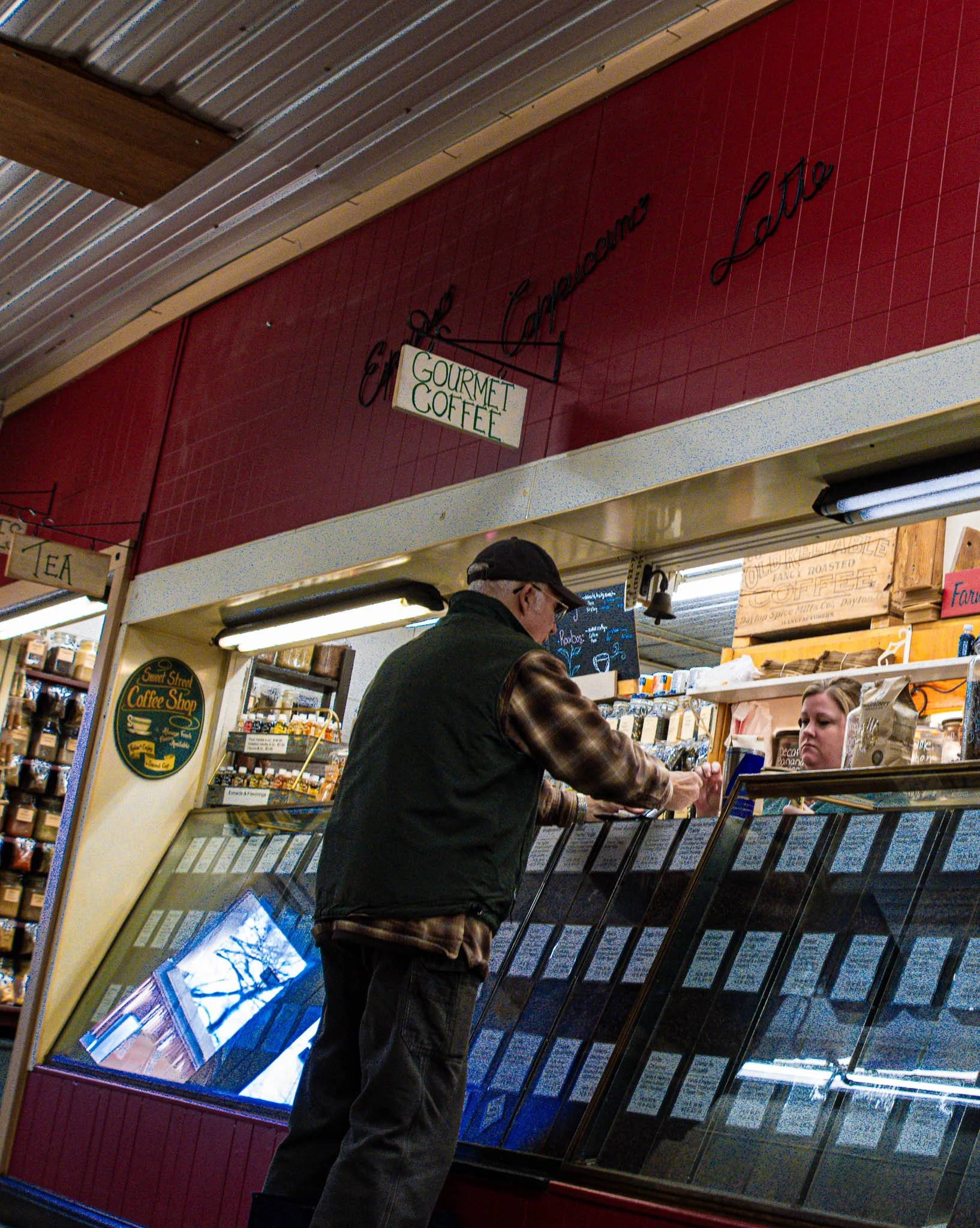 Man in dark vest and plaid shirt buying coffee from a glass display case in a cafe.