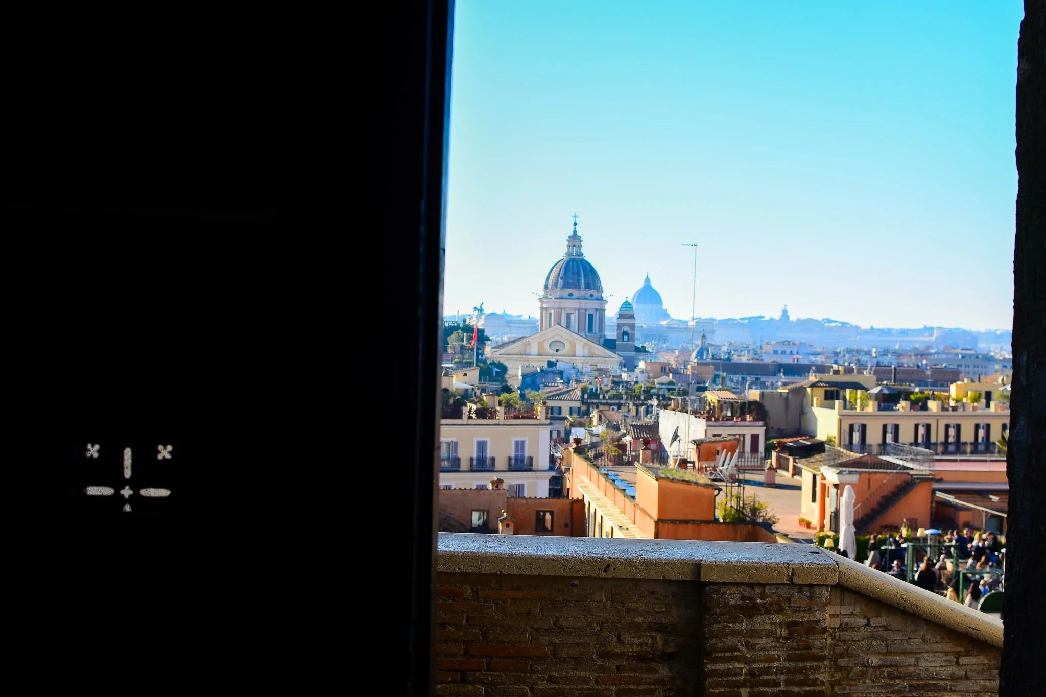 View of Rome's cityscape with a large dome and cathedral in the distance, seen through an opening or window, with blue sky overhead.