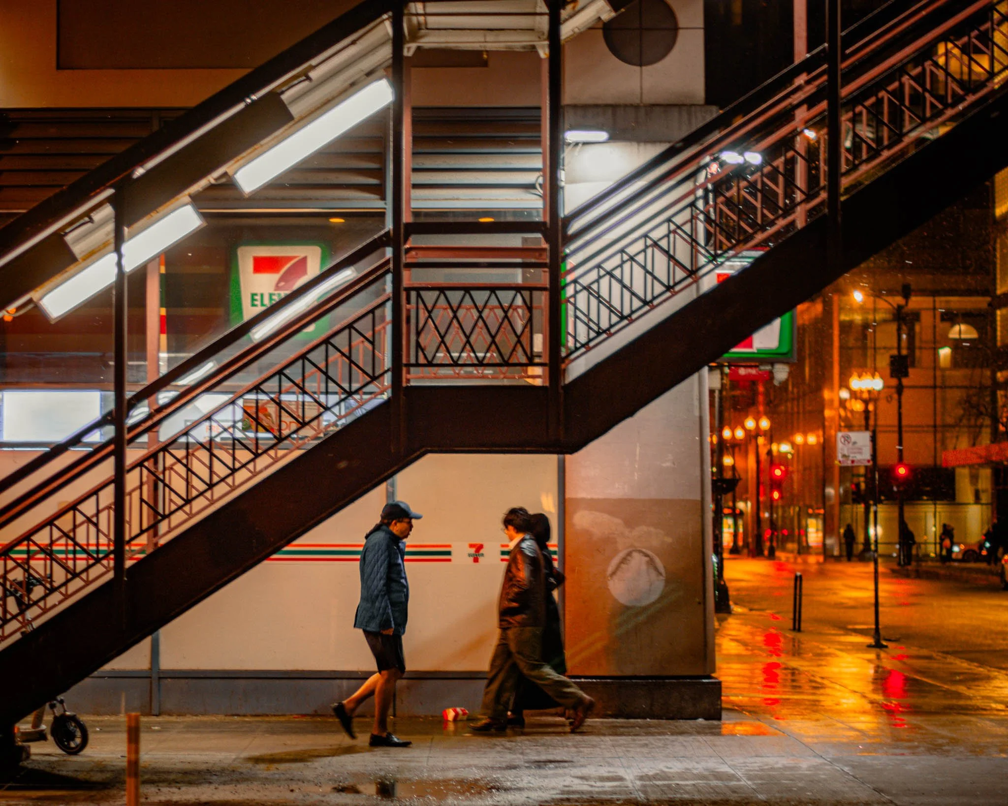 Two men walking near a staircase outside of a 7-Eleven store at night, with city street lights and reflections on wet pavement in the background.