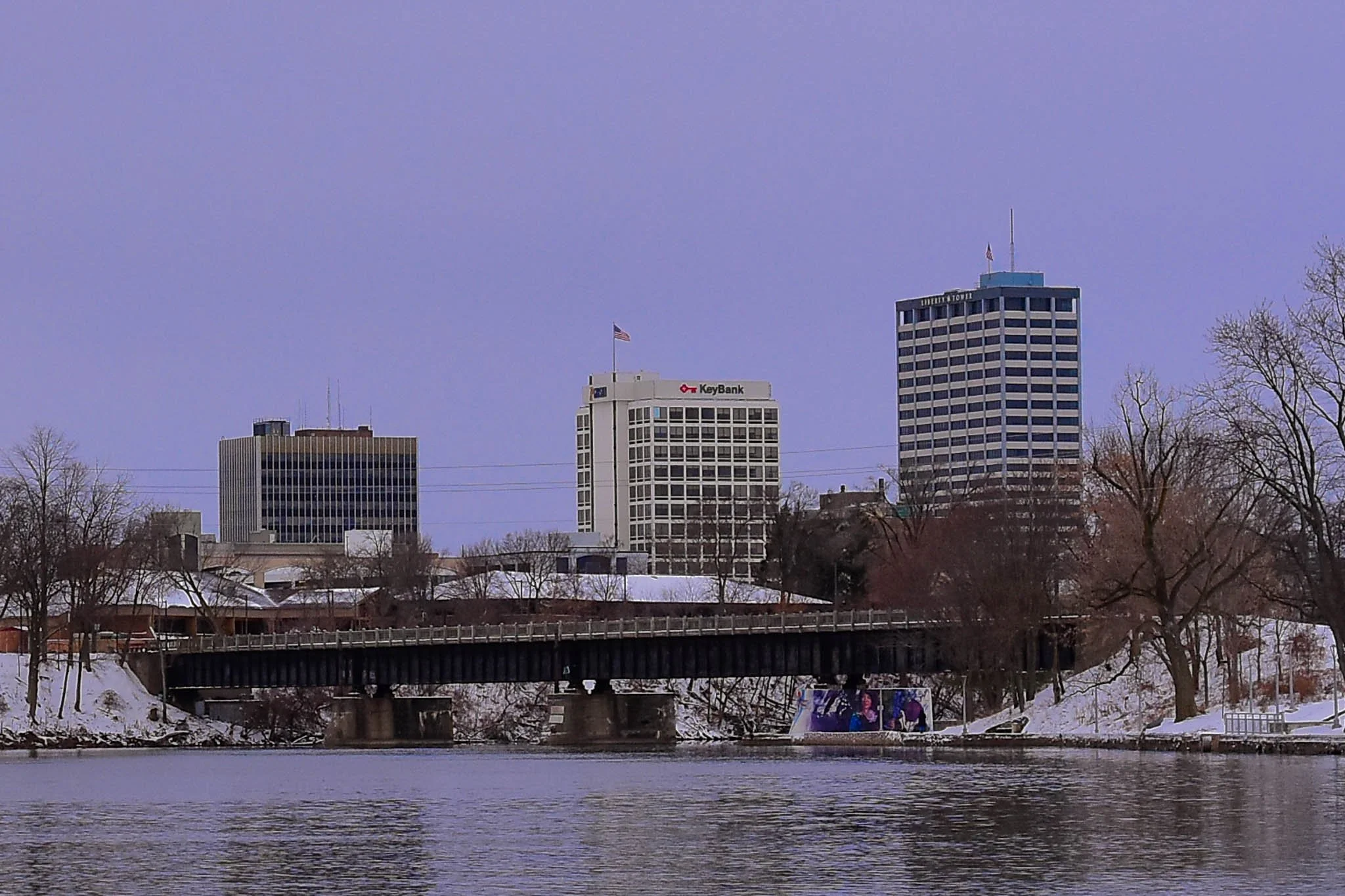 City skyline with tall office buildings, a bridge over a river, and snow-covered trees in the foreground, under a light purple sky.