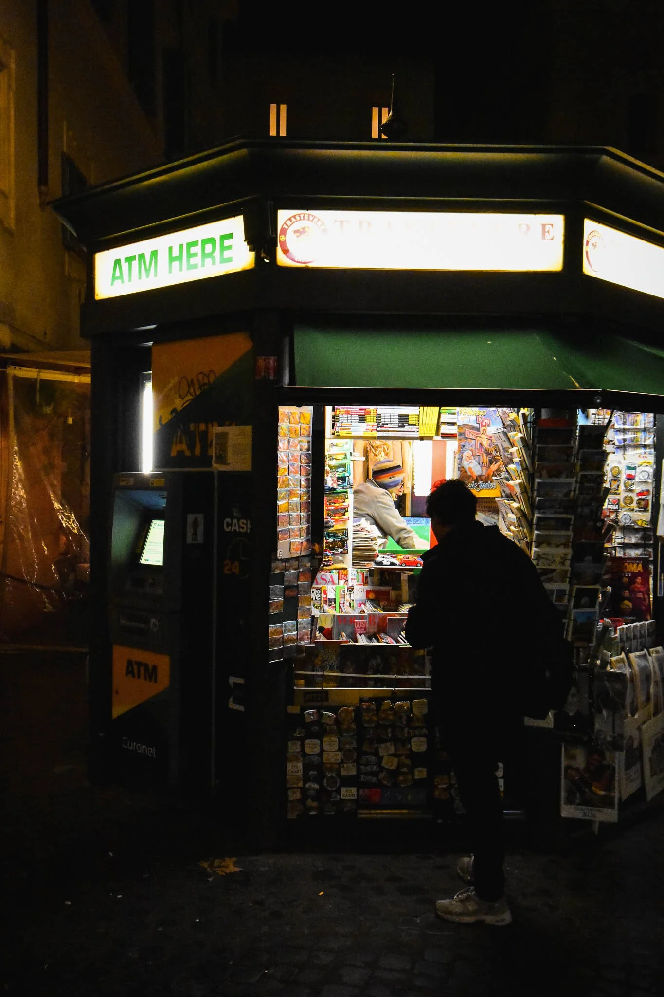 A small, illuminated newsstand at night with a green awning, selling magazines, souvenirs, and snacks. A person is standing in front of it, looking at the items inside, with a bright sign that reads 'ATM HERE' on top.