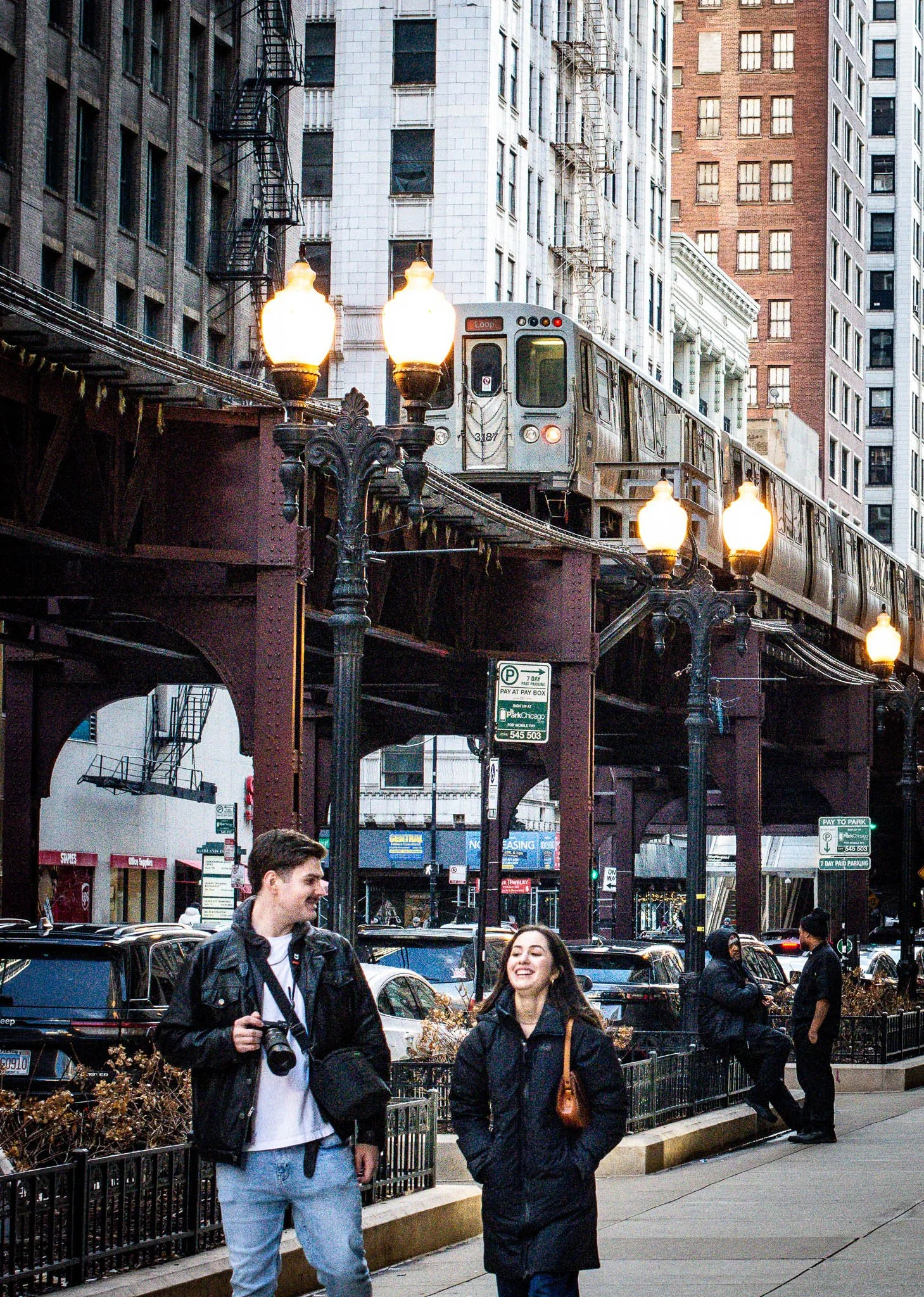 Street scene in an urban area with a train on elevated tracks, street lamps, parked cars, and pedestrians, including two young people smiling and talking.