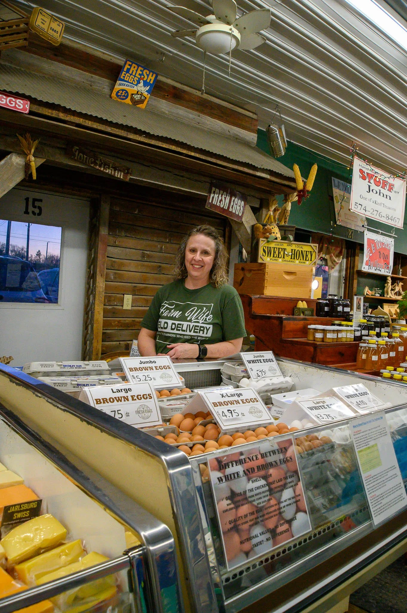 A woman behind a display of eggs at a farmers market stand, smiling, with signs indicating different types of eggs and prices, surrounded by other market items and signs.