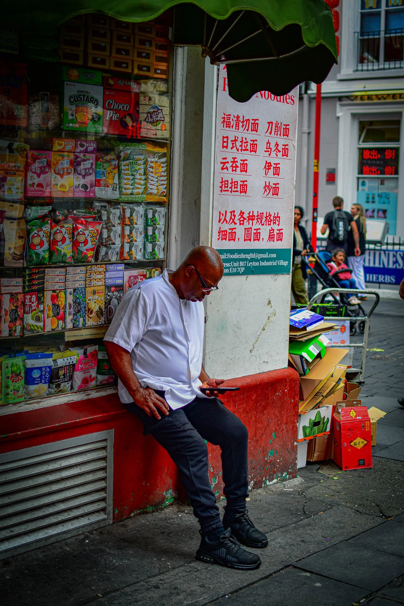 A bald man with glasses wearing a white shirt and black pants sitting outside a shop, looking at his phone. The shop window displays various colorful snack packages. There's a sign with Chinese characters and an email address on the wall behind him. 