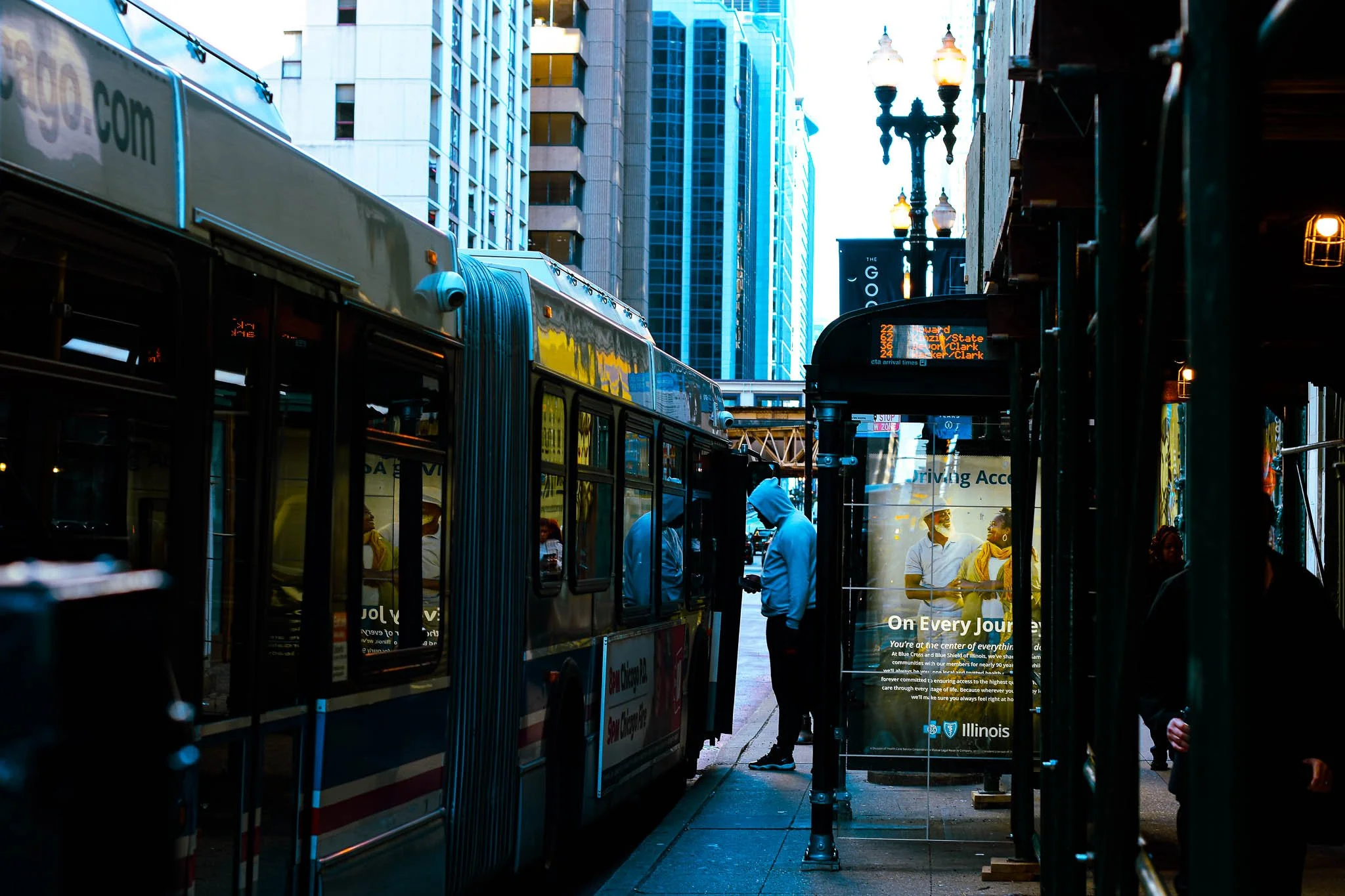 A city street scene with a bus at a bus stop and people waiting. Tall buildings and a digital bus schedule display are visible.