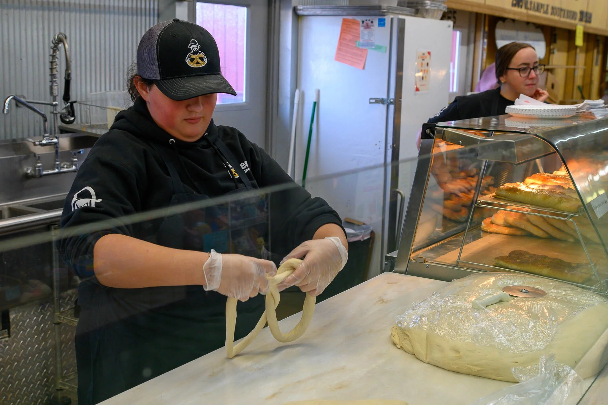 Person with black baseball cap and black hoodie preparing dough in a bakery or pizzeria, with a display case of baked bread or pastries nearby.