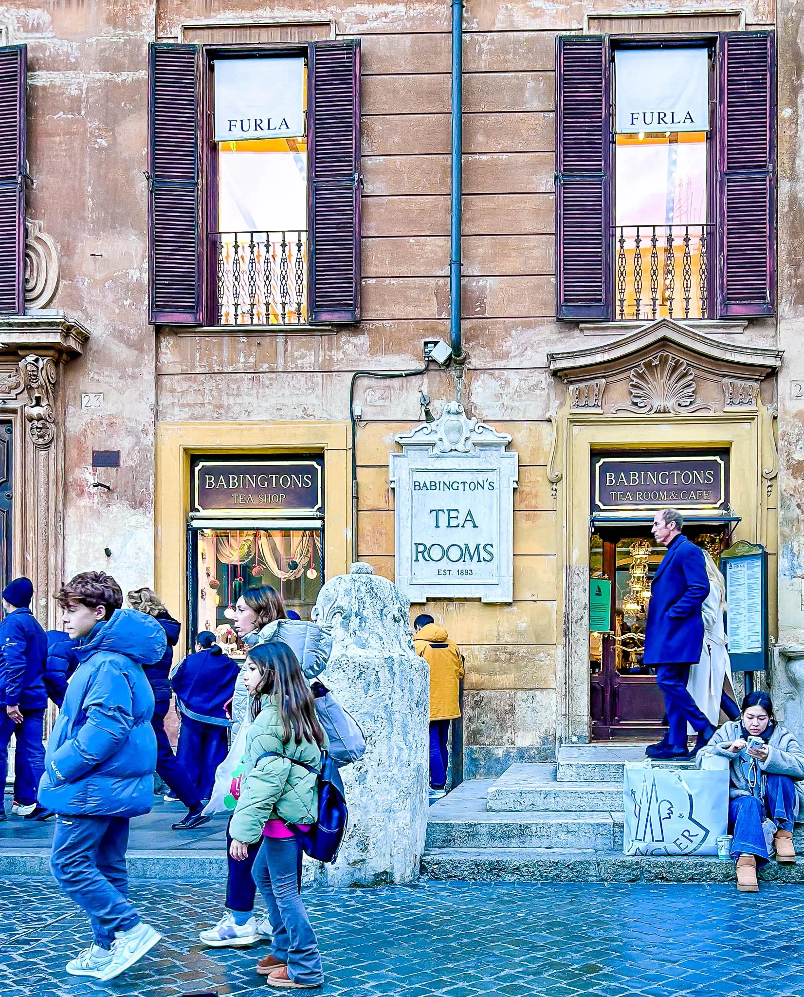 The photo shows a street scene in front of Babington's Tea Room & Cafe, with people walking and sitting outside. The building has a rustic facade with purple window shutters, and signs for Babington's Tea Room and Tea Shop. There is a marble fountain