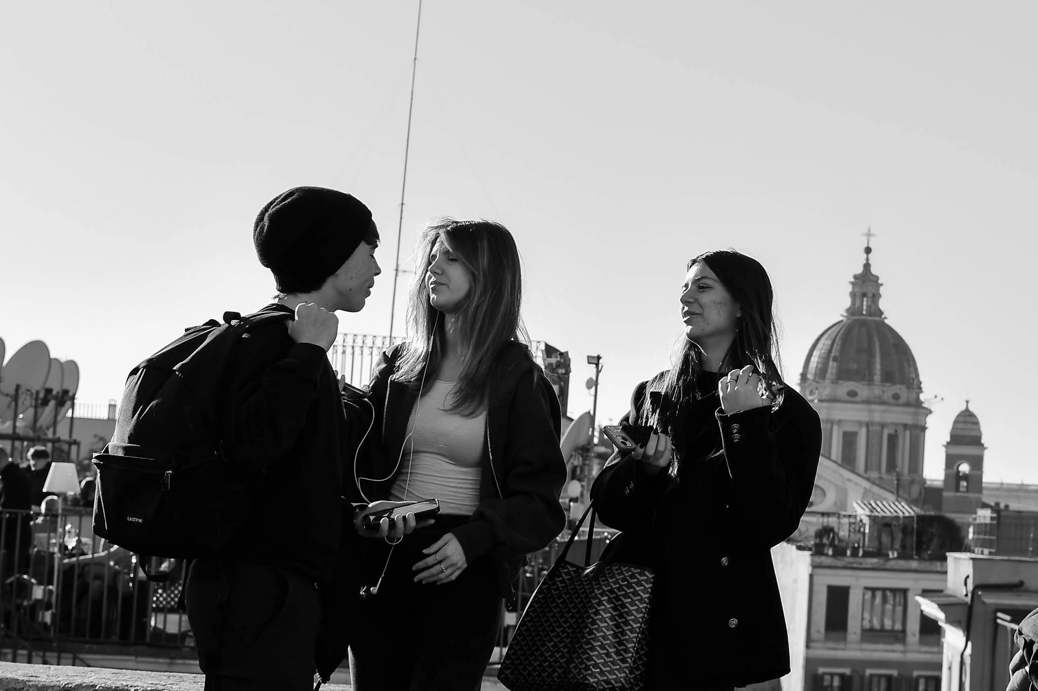 Three young women are standing outdoors on a city rooftop or terrace with a historical building featuring a large dome in the background. They are engaged in a conversation, holding smartphones, with one of them wearing headphones. The scene is in bl