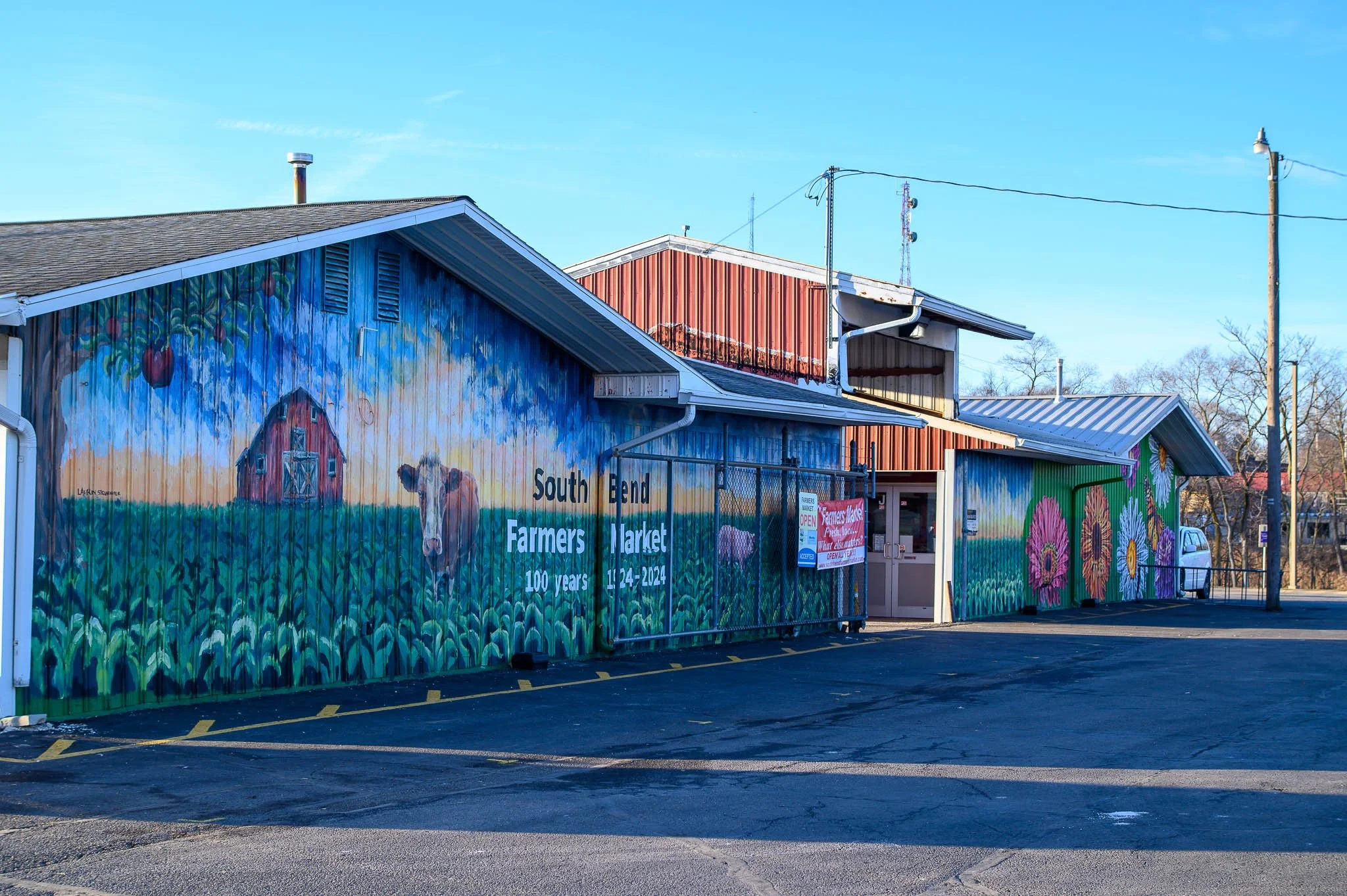 Colorful mural on a building advertising the South End Farmers Market, including a barn, cow, and large flowers, with signage indicating it is celebrating 100 years from 1924 to 2024.