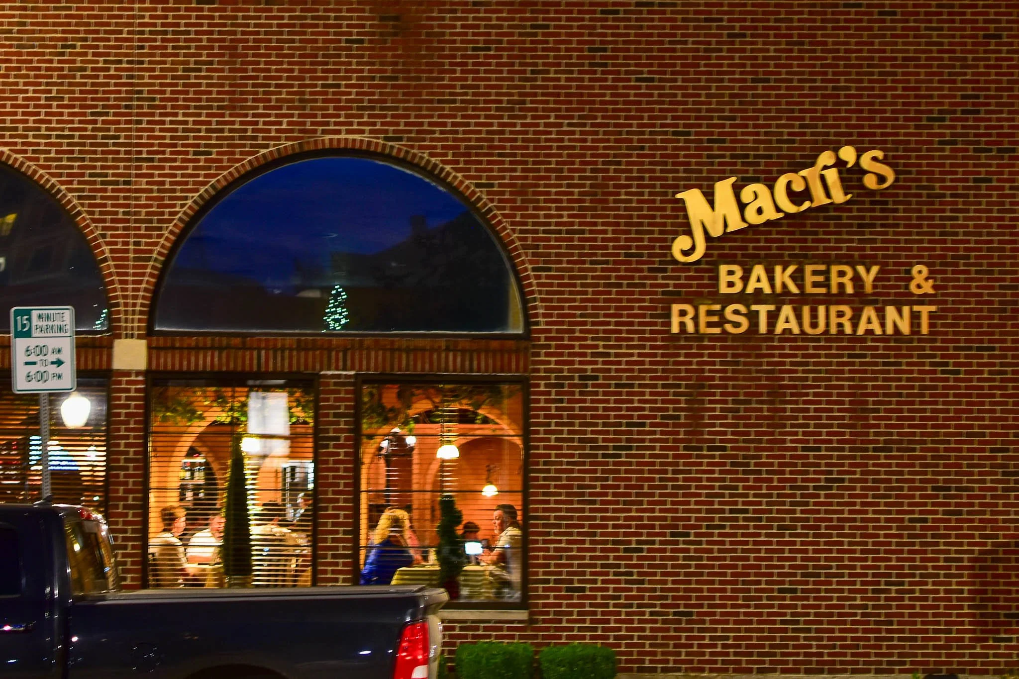 The exterior of Macit's Bakery & Restaurant with a brick wall, large arched windows showing people dining inside, and a sign on the wall. A parking sign is also visible.