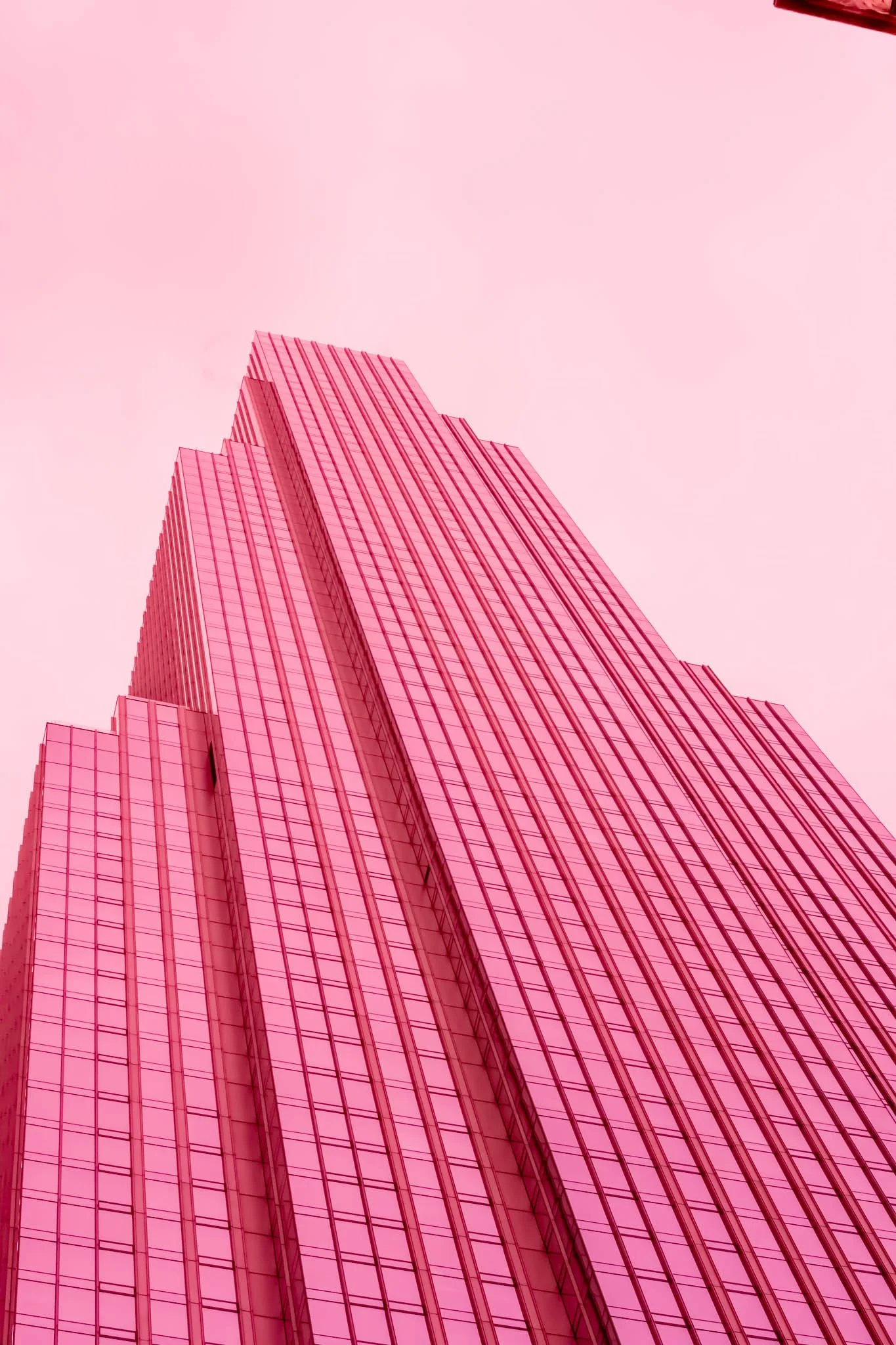 Pink skyscraper with glass windows seen from below against a pink sky.