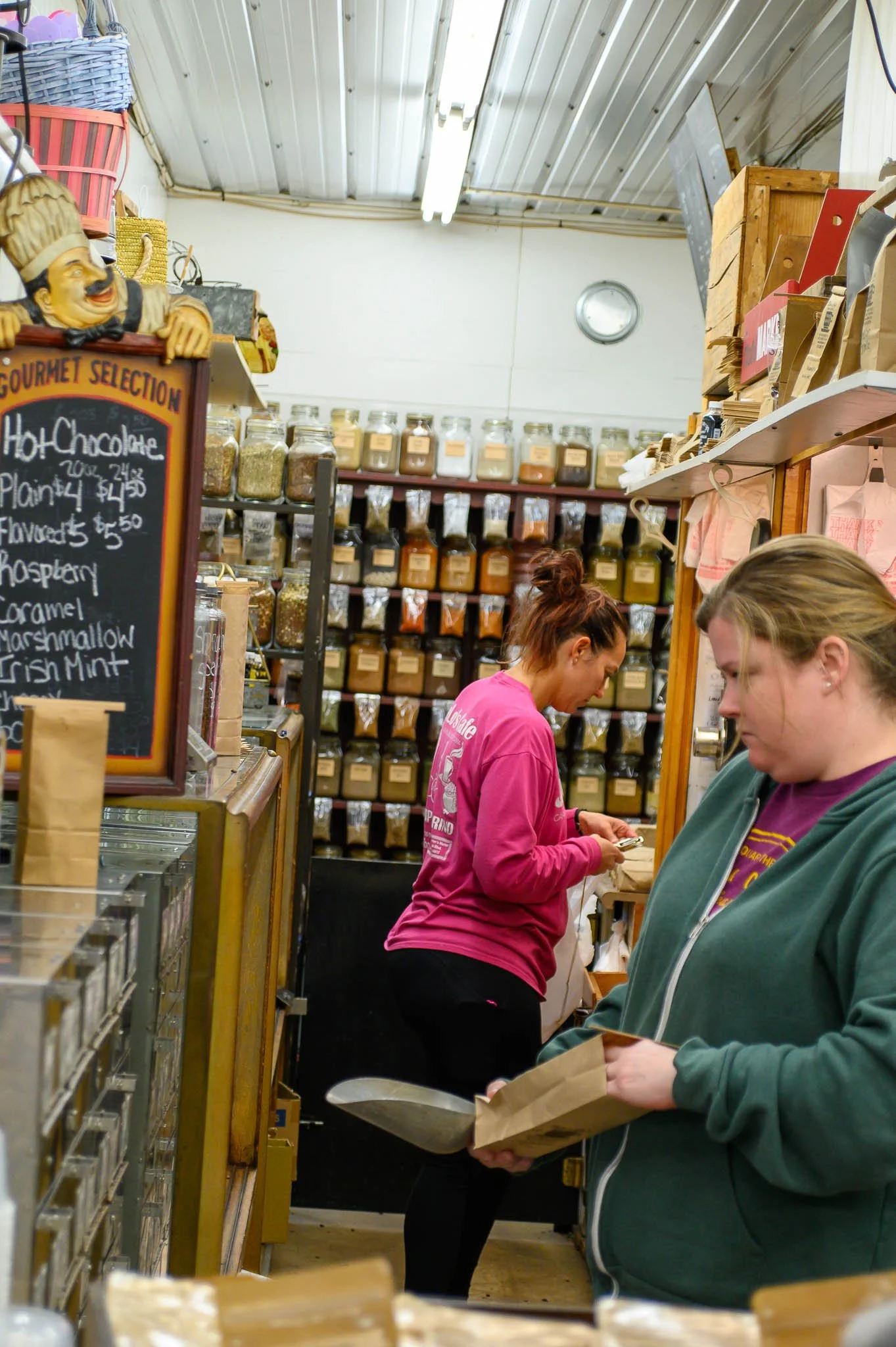 Two women shopping in a bulk food store with shelves of labeled jars and bags of spices and dry goods in the background.