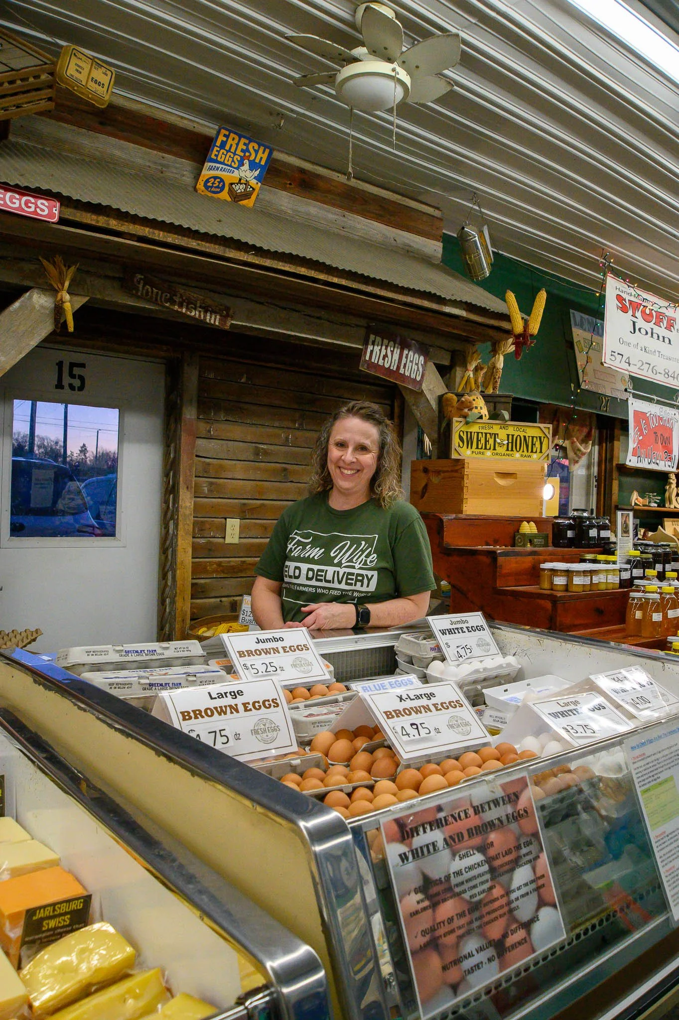 A woman shopkeeper standing behind a display of brown, blue, and white eggs at a farm stand. Signs display prices, and the background includes wooden decor, honey jars, and signs advertising fresh eggs and local honey.