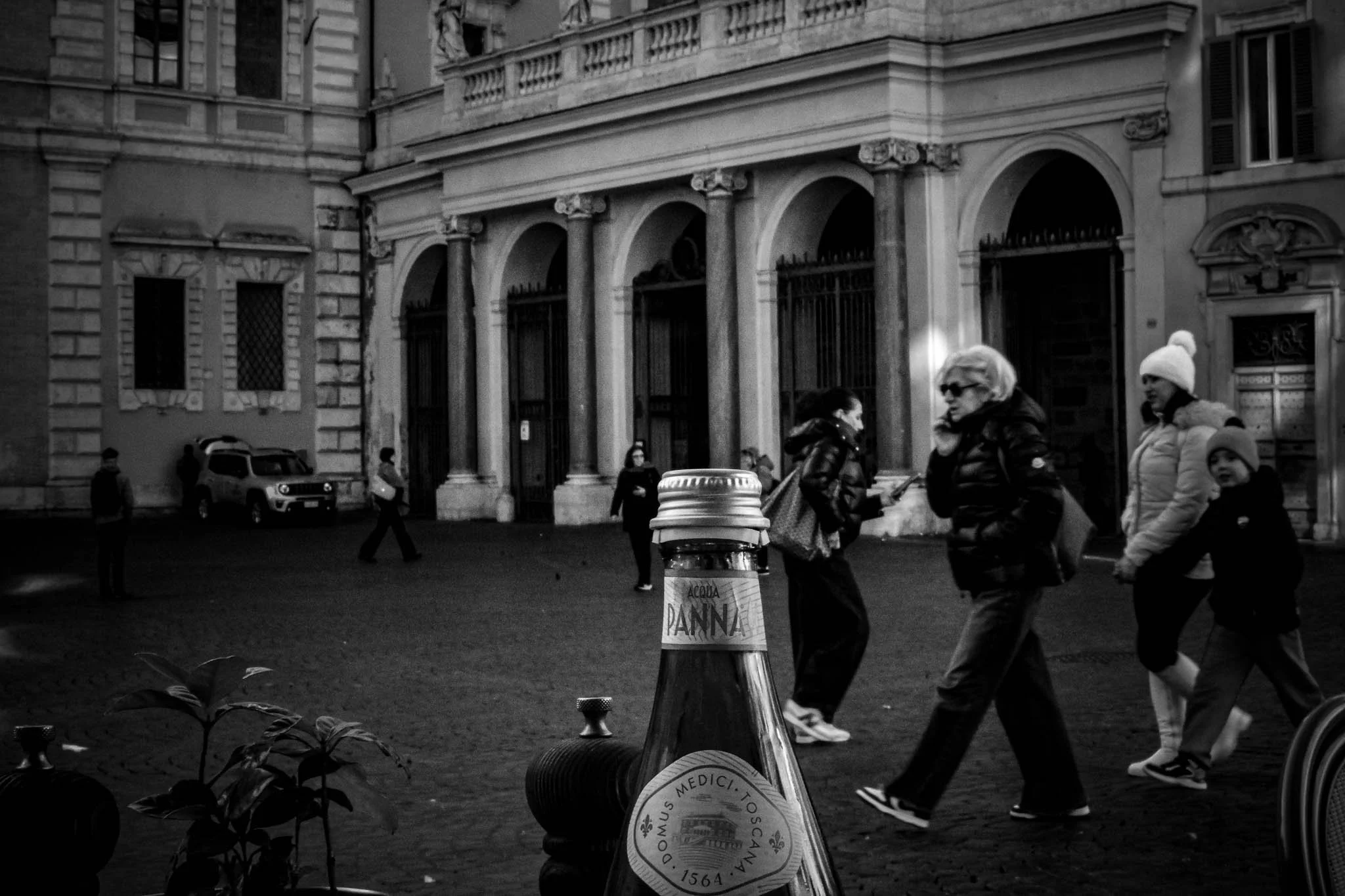 Street scene in black and white with pedestrians walking past historic buildings, and a bottle of water labeled 'Acqua Panna' in the foreground.