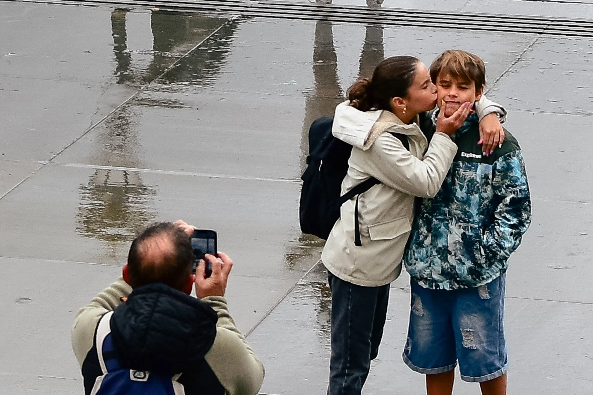 A woman is pecking a boy on the cheek, both are standing on a rainy sidewalk. A man is taking a photo of them with his phone.
