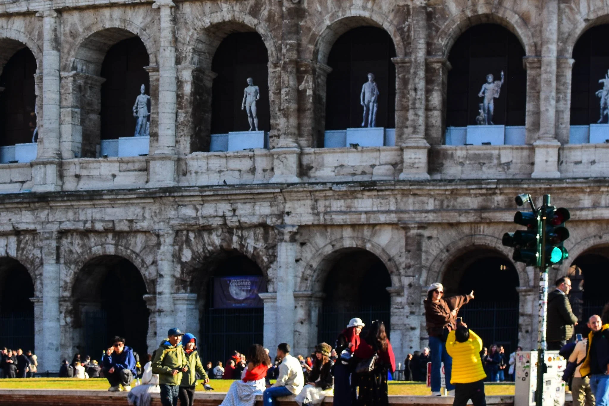 People gather in front of the Colosseum in Rome, Italy, with some tourists taking photos and others sitting on the grass. The historic structure's arches and ancient stone facade are visible, along with several statues in the upper arches.