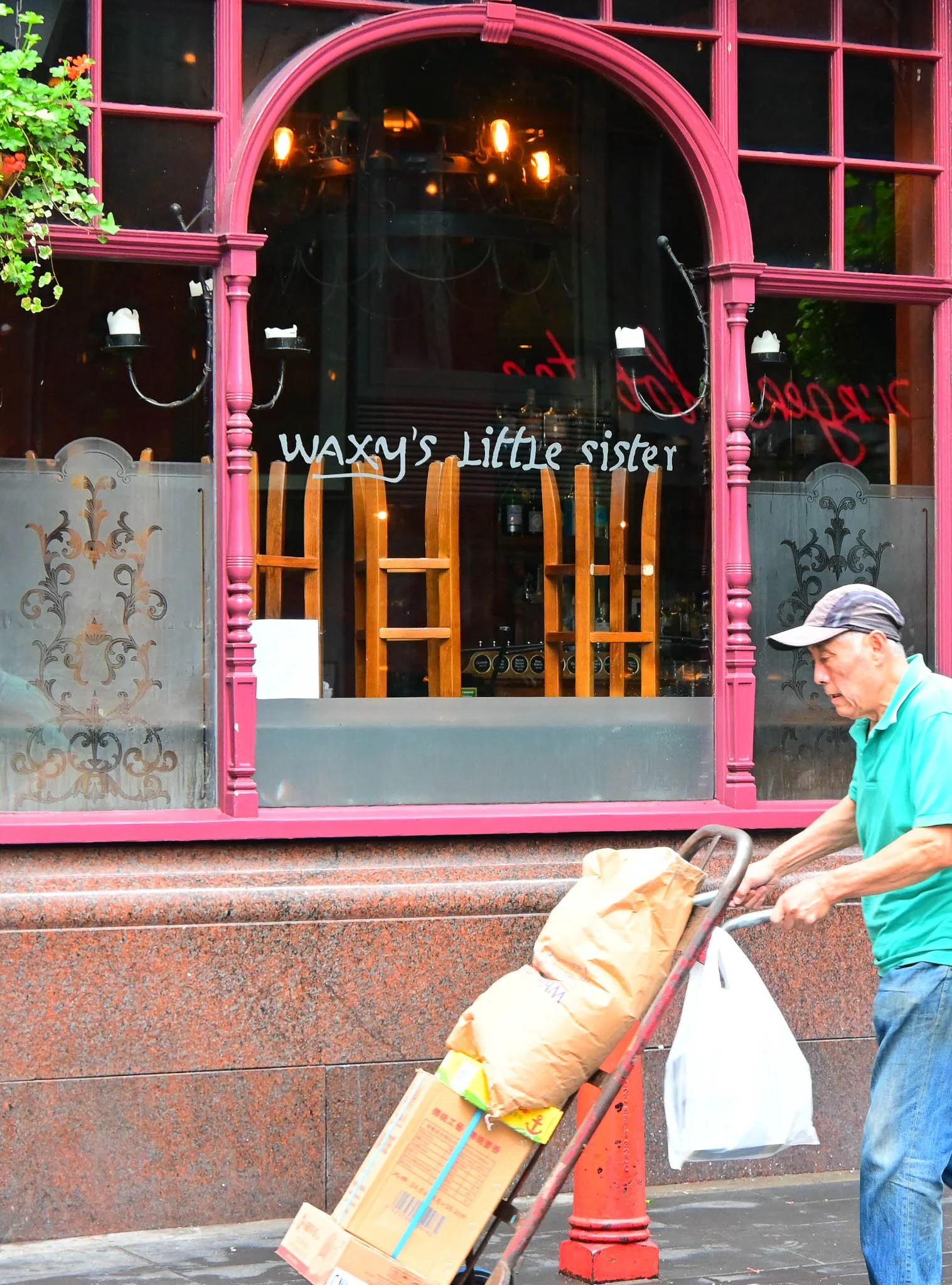 A man wearing a gray cap, teal polo shirt, and jeans pushes a hand truck loaded with a large paper bag and a flattened cardboard box past a restaurant window with pink framing that reads 'Waxy's Little Sister' in white letters.