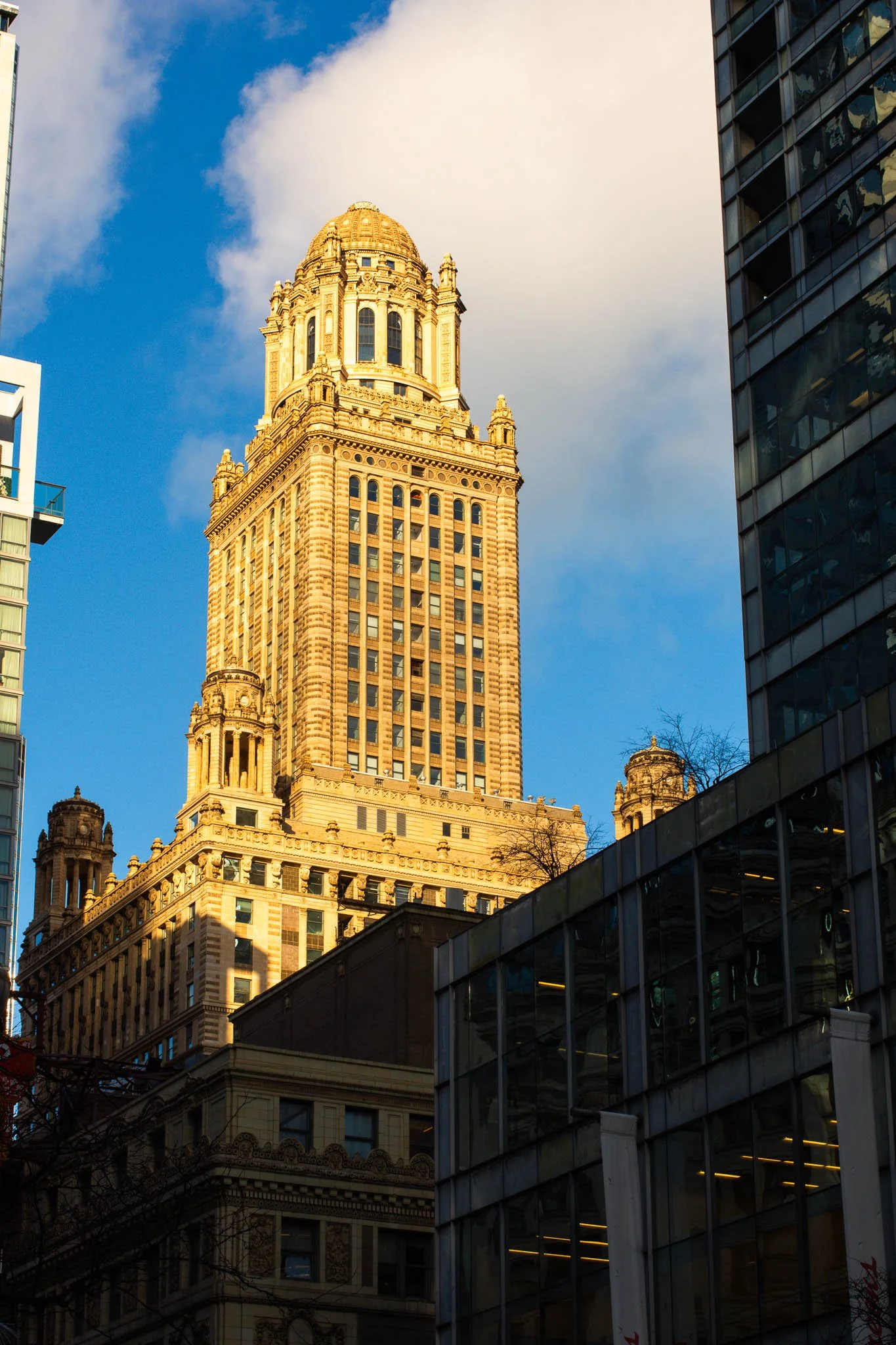 A tall, ornate skyscraper in a city skyline, with a decorative dome at the top, surrounded by other modern buildings, under a blue sky with some clouds.