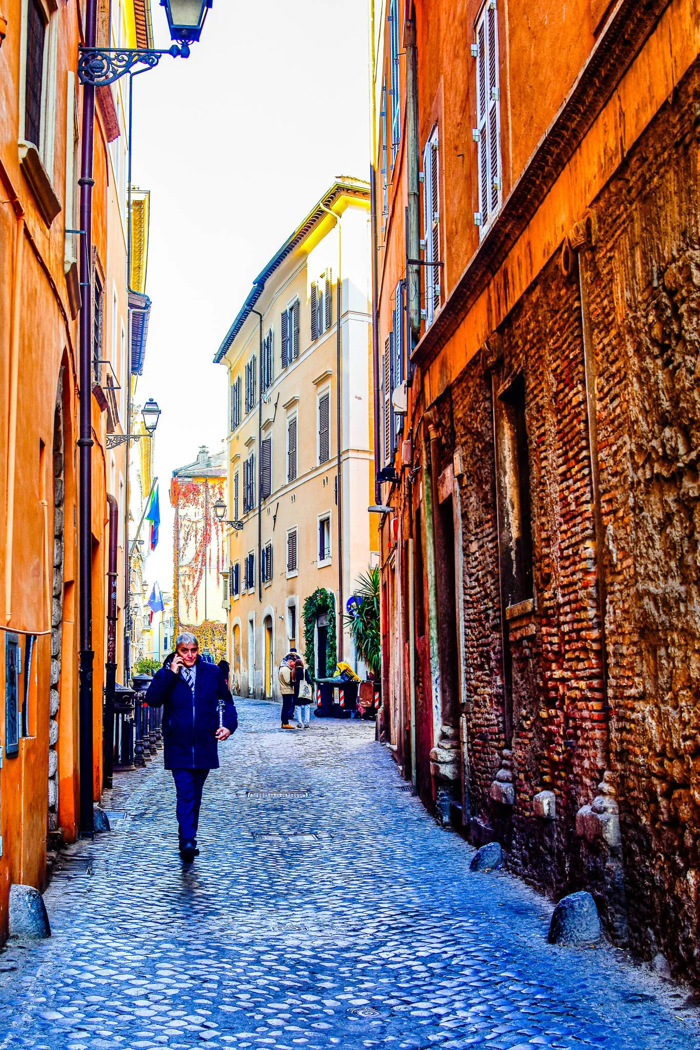 Narrow cobblestone street in a European city, lined with colorful buildings and a man wearing a dark coat walking while talking on his phone.