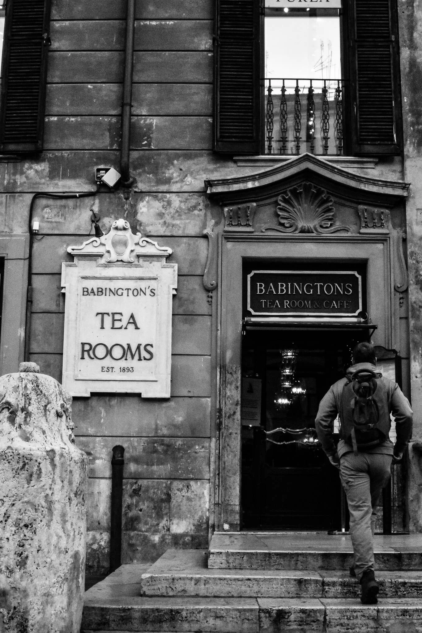 A person with a backpack walking into Babington's Tea Room & Cafe, established in 1893, with signs and an ornate doorway.