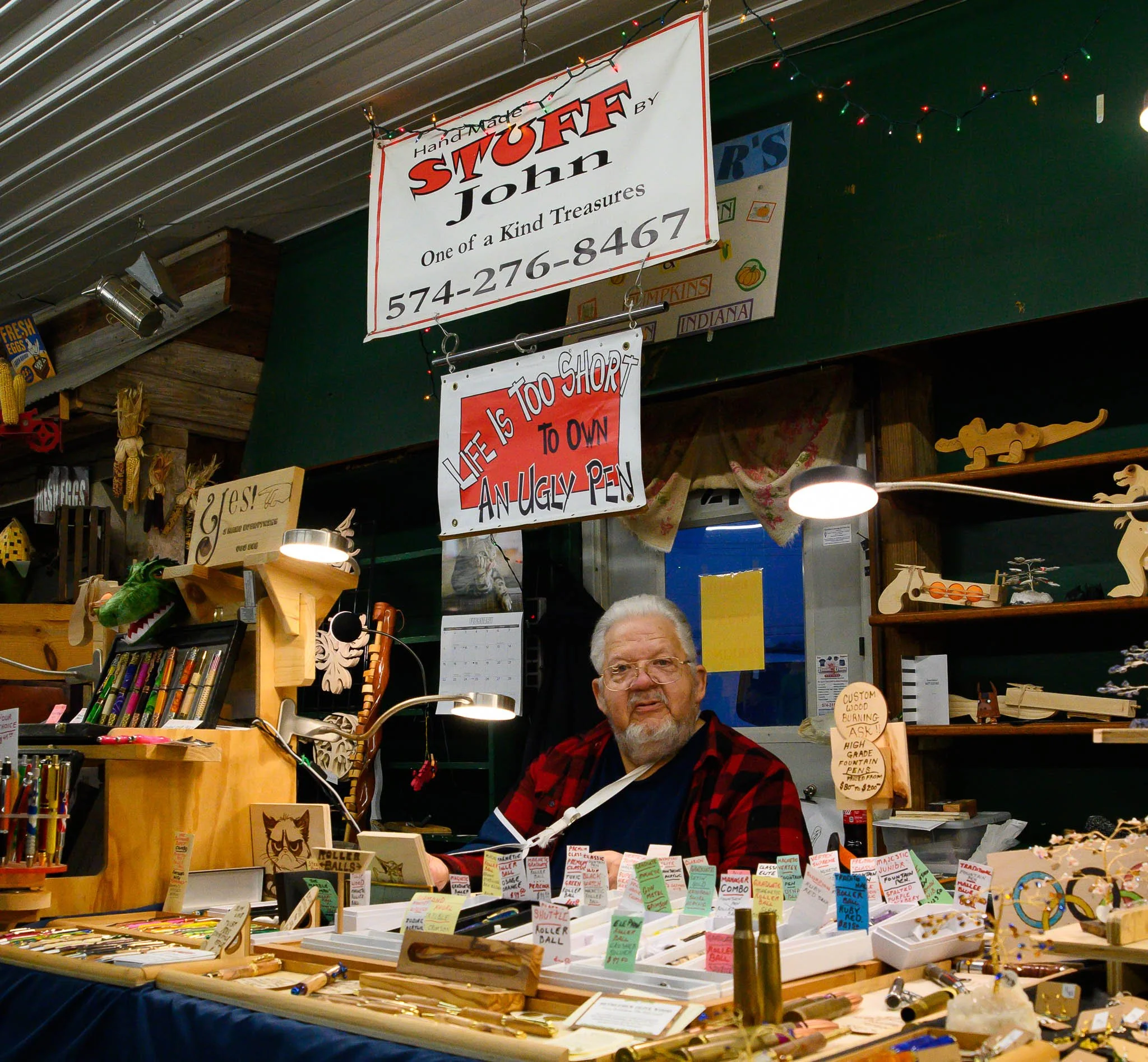An elderly man with gray hair and glasses sits behind a display table filled with pens and woodworking items at a craft fair. Signs above his booth include one that reads 'Handmade by StuFF John' and a humorous sign stating 'Life is too short to own 