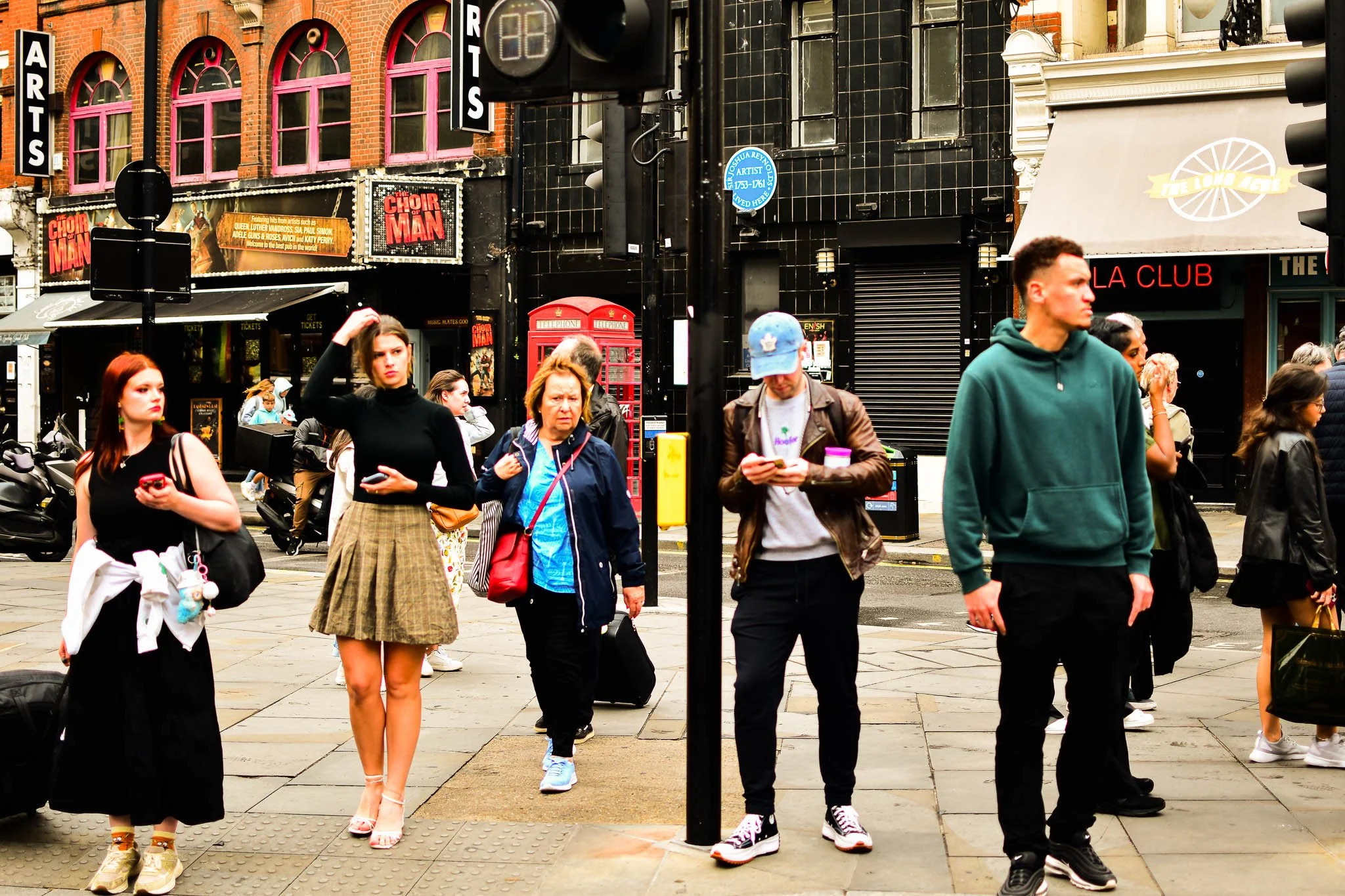 People waiting at a city crosswalk, some looking at their phones, in front of a theater with show signs, a red phone booth, and storefronts.