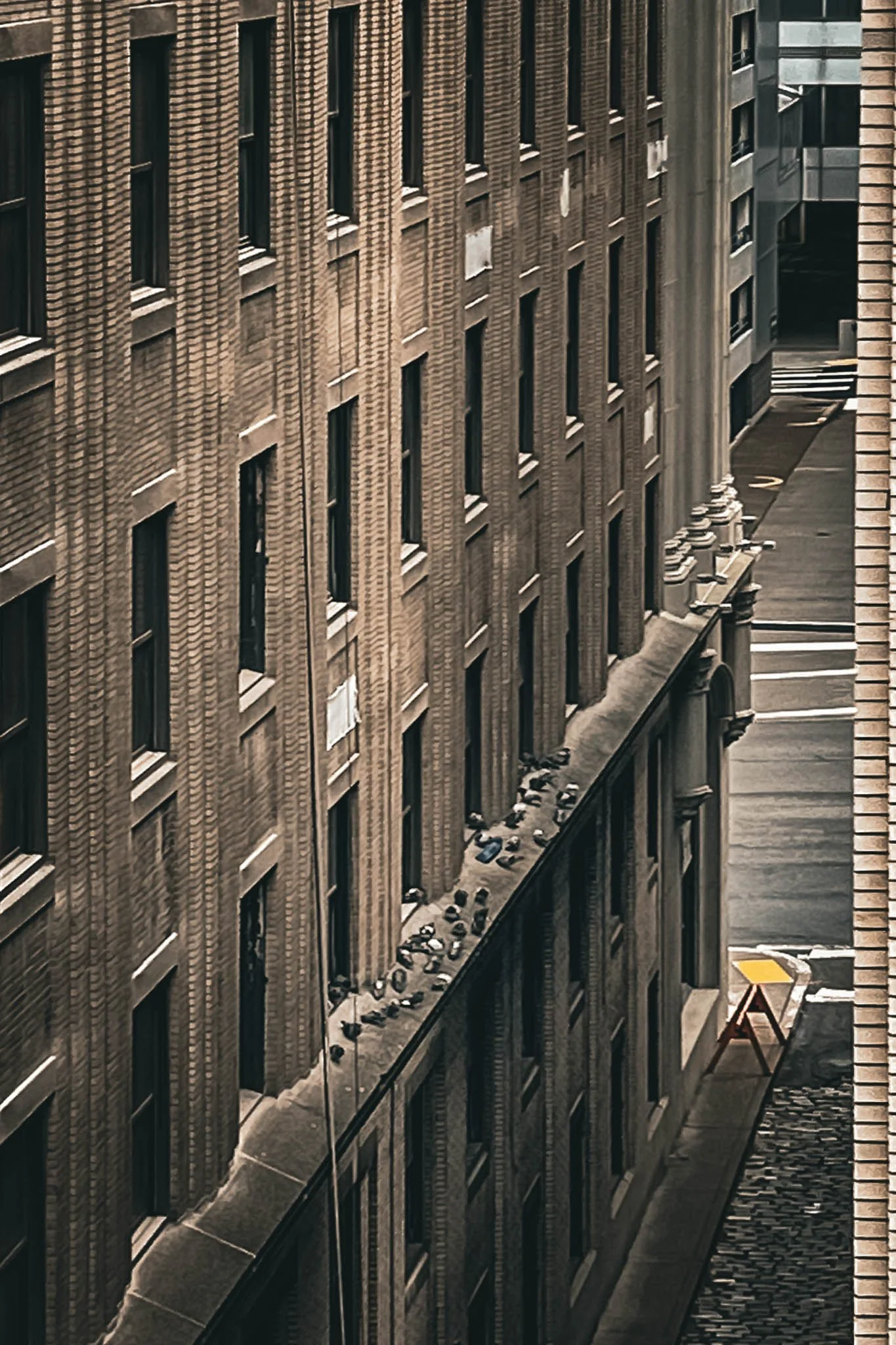 View of a tall brick building with multiple windows, some with air conditioning units, and what appears to be a narrow alley or parking area between buildings with scattered debris or objects on the ground.
